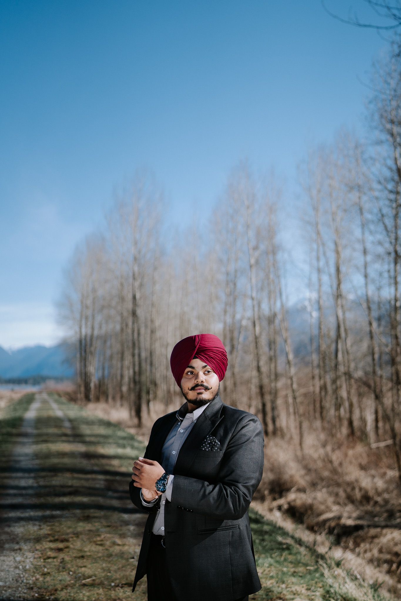 A person in a dark suit and burgundy turban stands on a rural path with bare trees and mountains in the background.
