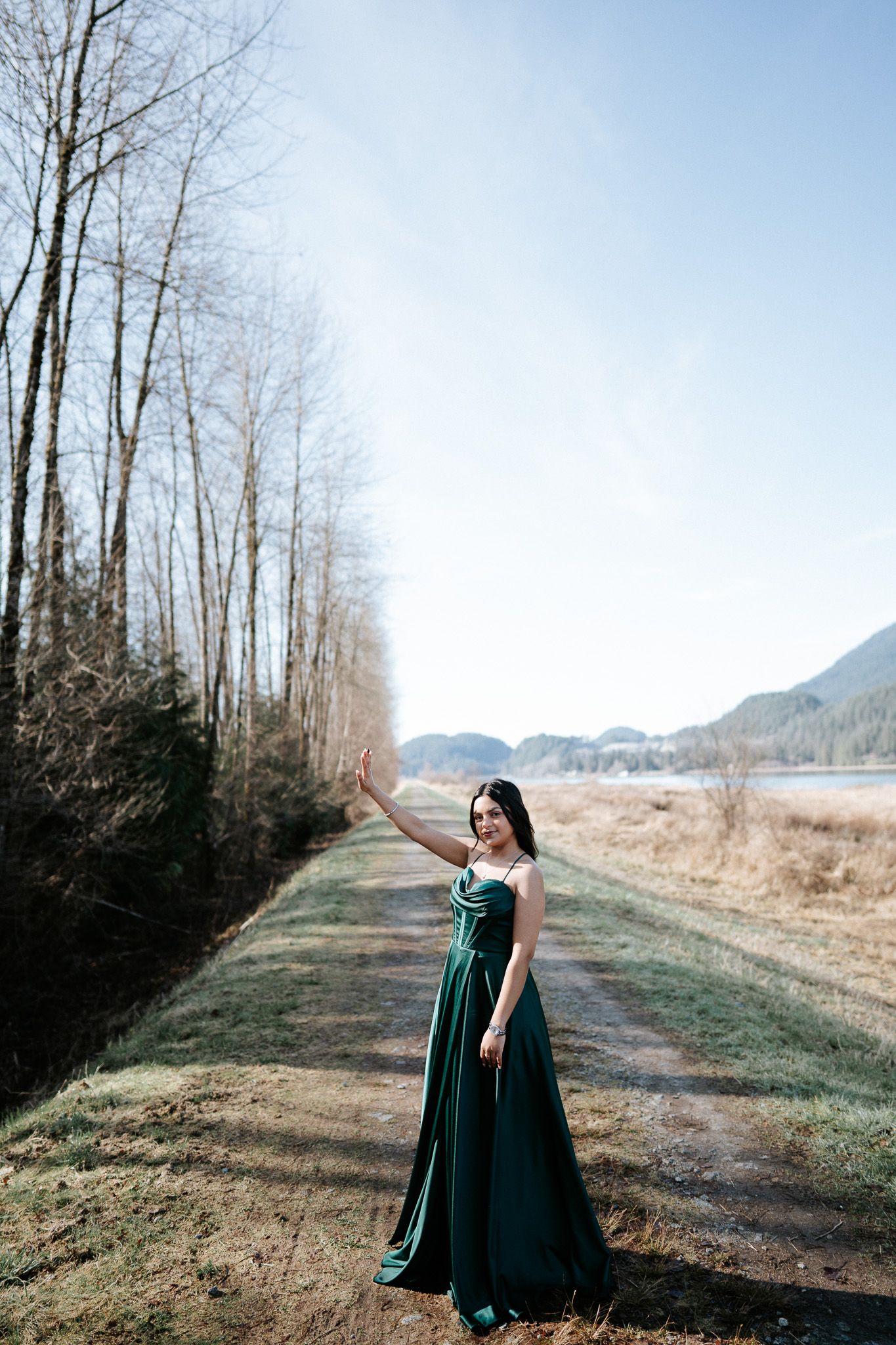 A figure in a flowing emerald green dress stands on a misty rural path bordered by bare winter trees and mountains.