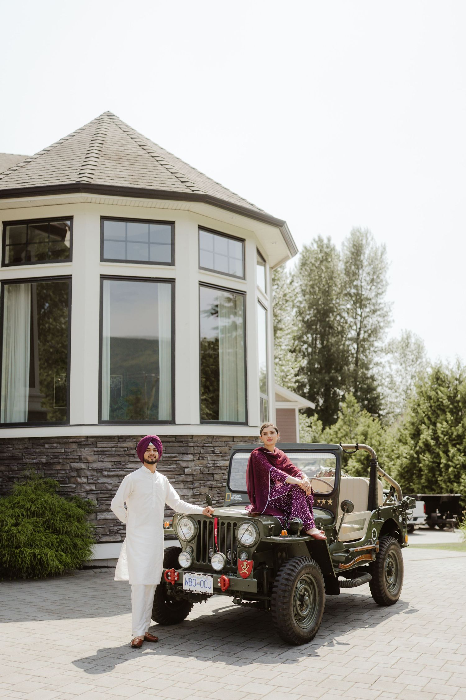 Sikh couple posing with a vintage military jeep outside a stone building, wearing traditional Punjabi attire.