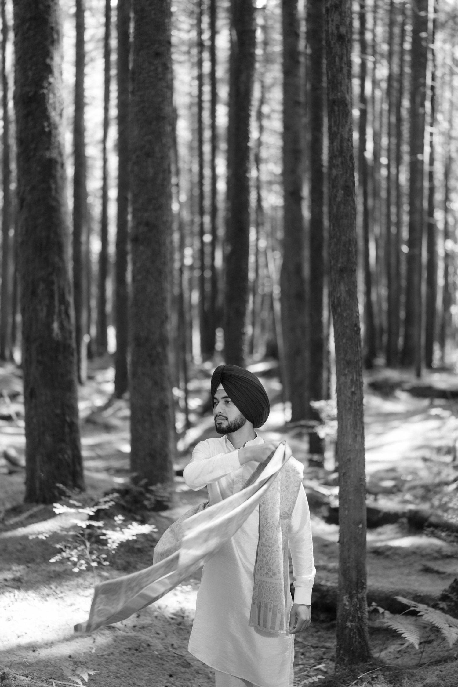 Black and white portrait of a Sikh man in white kurta and turban standing in a pine forest.
