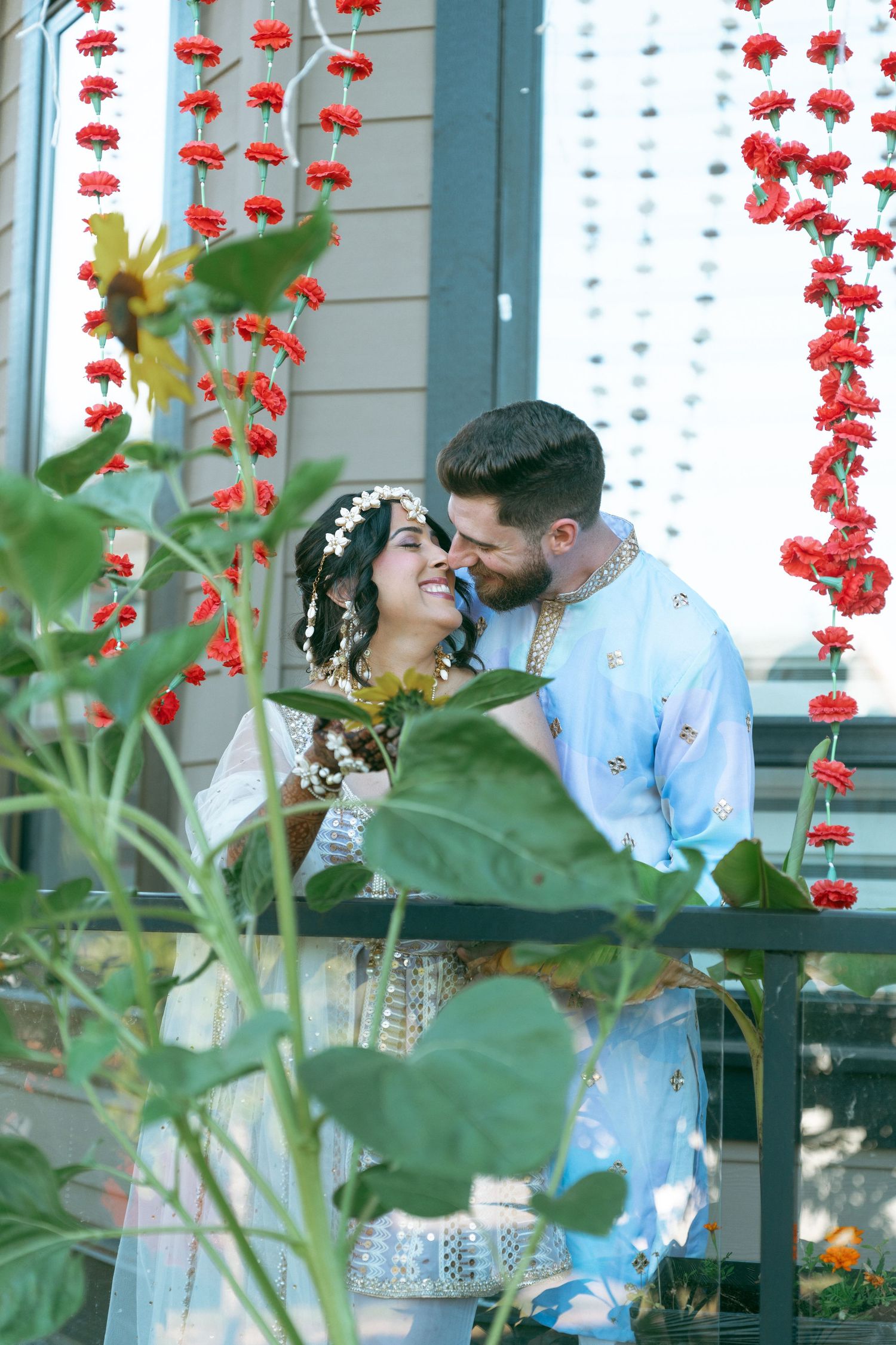Couple sharing a romantic moment on a balcony adorned with red floral garlands during a traditional wedding ceremony.
