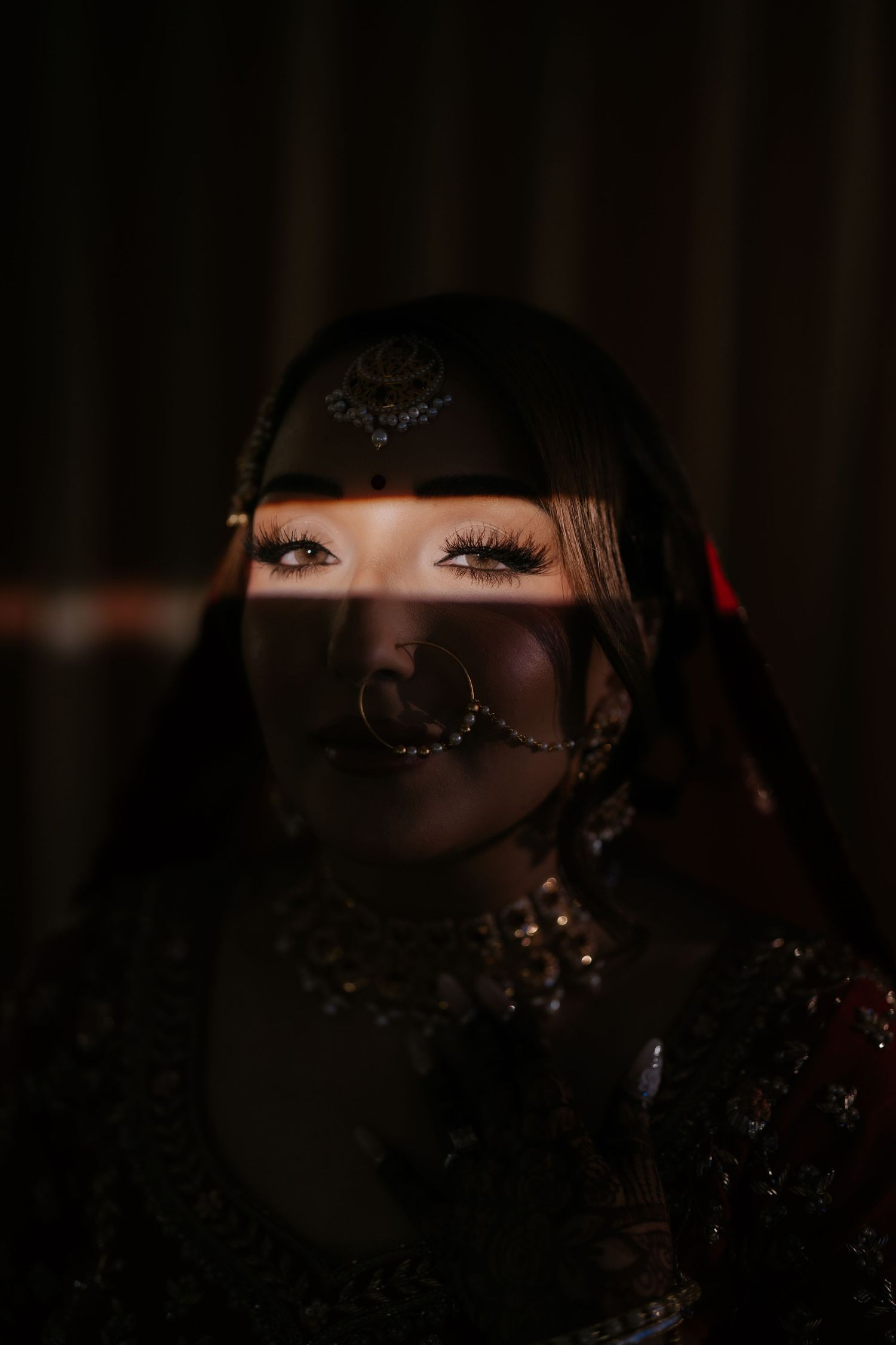 Woman in traditional Indian bridal jewelry with maang tikka and nose ring, dramatically lit against dark background.