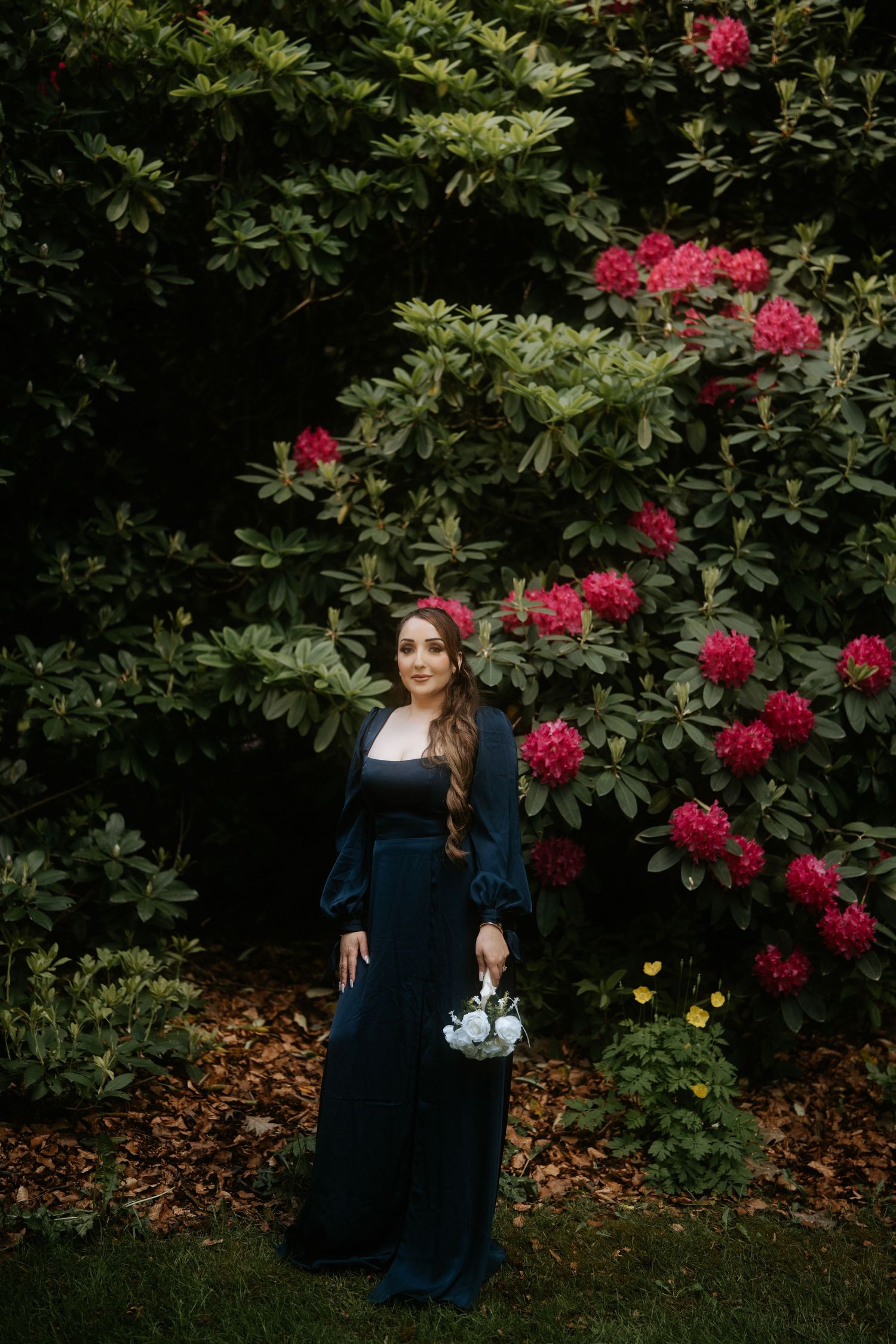 Woman in dark gown holding white flowers stands before blooming pink rhododendrons in a lush garden.