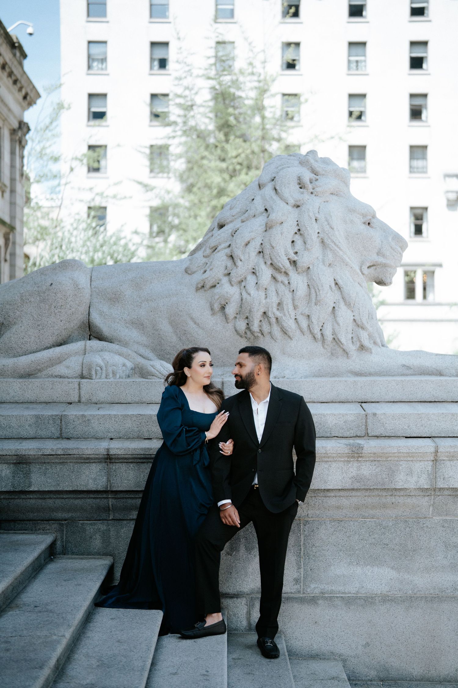 Couple poses by a stone lion sculpture outside a grand building, woman in navy gown, man in black suit.