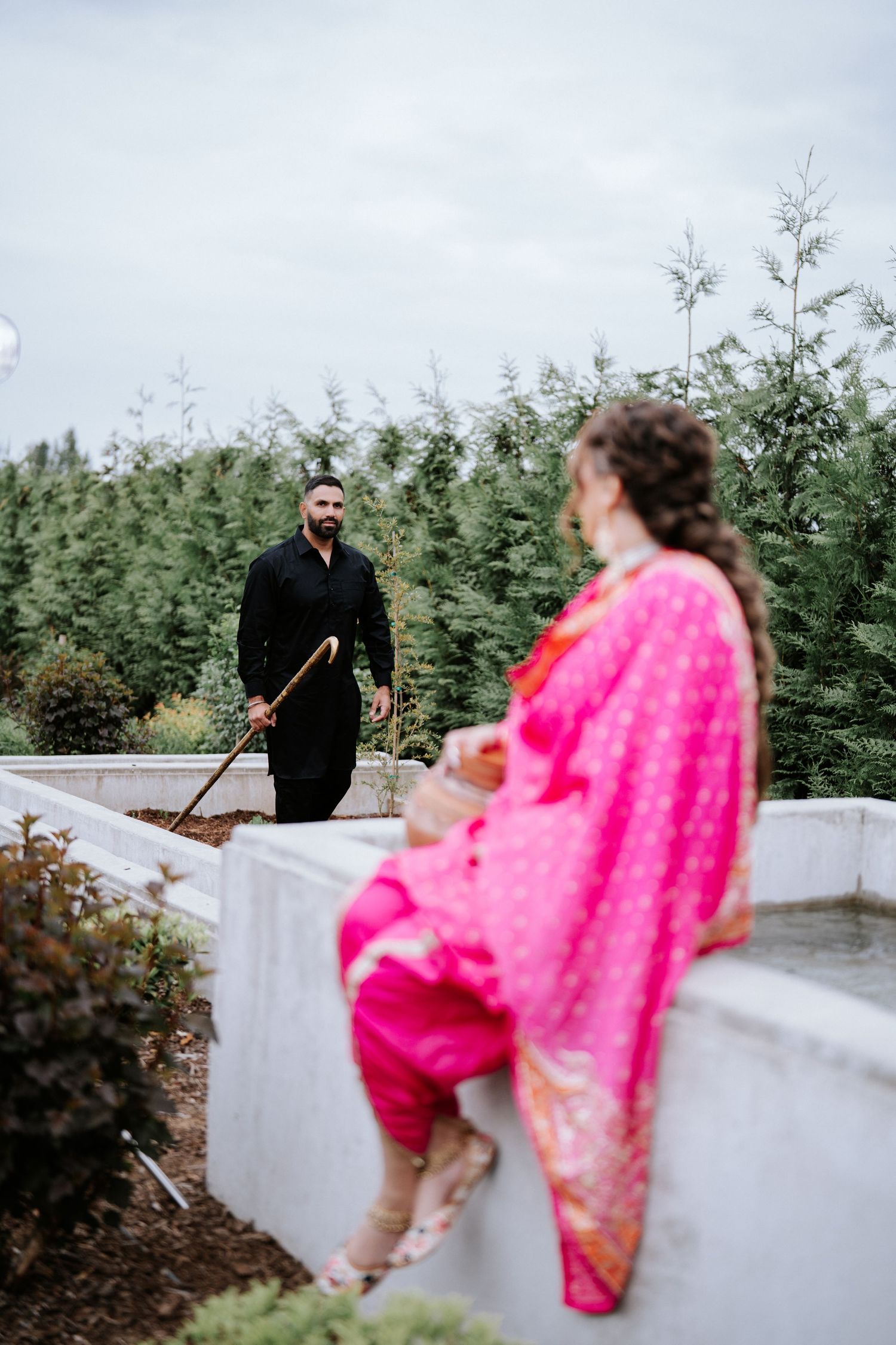 A man in black kurta holds a stick while a woman in pink salwar kameez sits on a white wall in a garden.