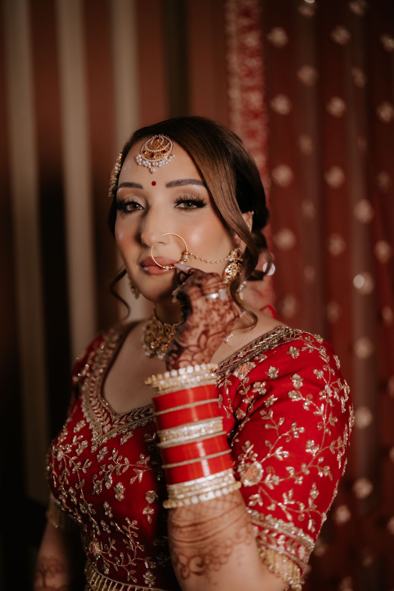 Indian bride in red lehenga with gold embroidery, wearing traditional bridal jewelry, maang tikka, and mehndi, posing elegantly.