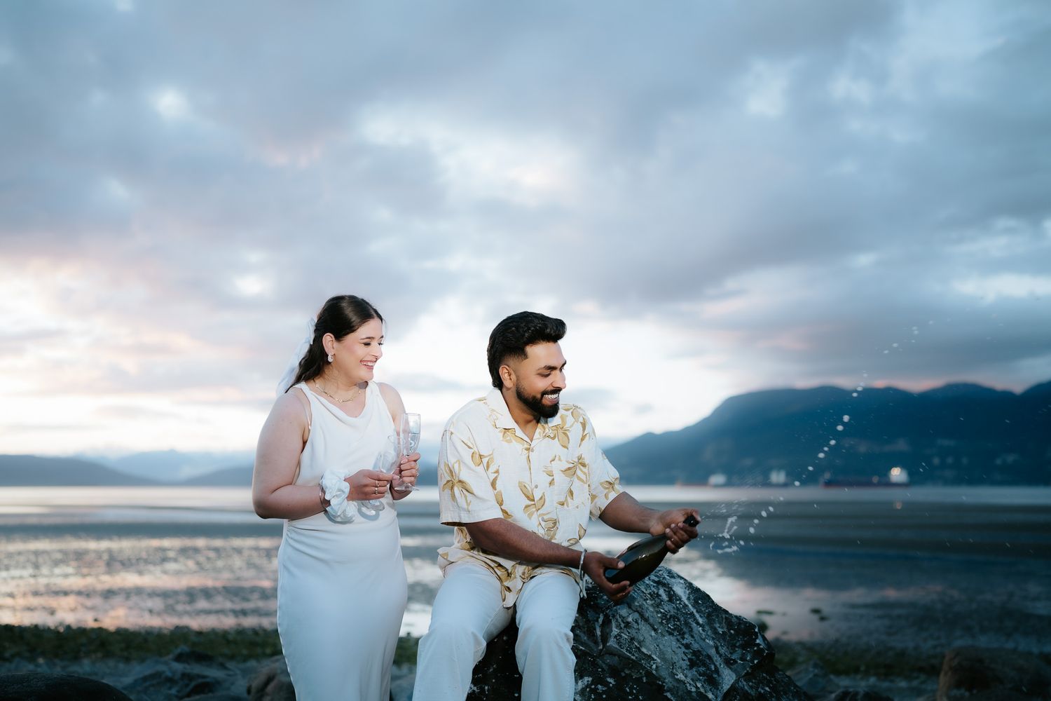 Couple celebrating outdoors by the ocean at sunset, woman in white dress and man in floral shirt popping champagne.