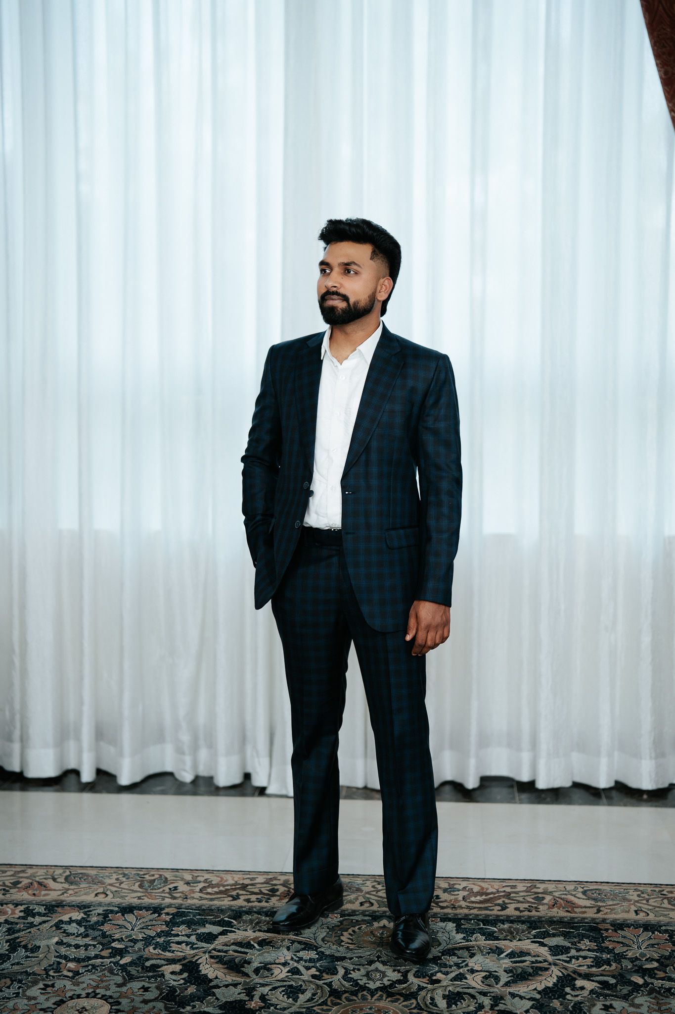 Stylish bearded man in dark plaid suit standing confidently before sheer white curtains on patterned rug.