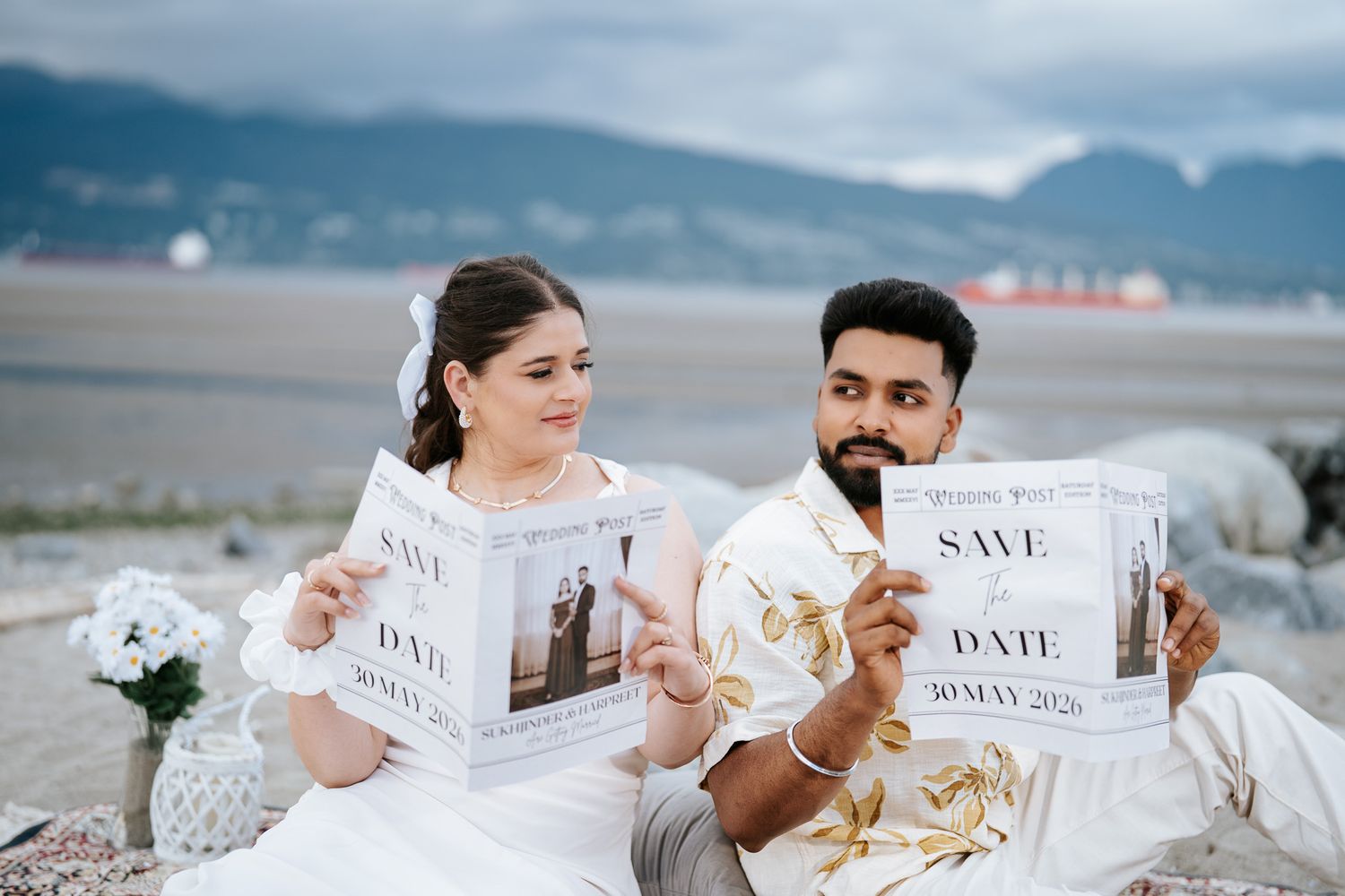 Couple sitting on beach holding Save the Date cards for May 30, 2026, with mountains in background.
