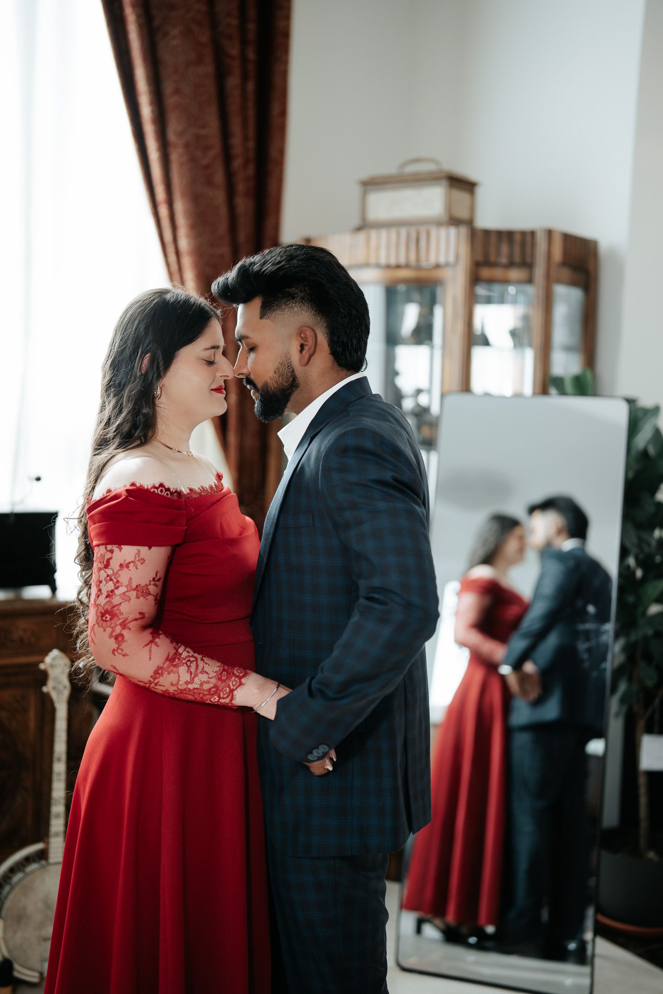 Couple in elegant attire sharing a romantic moment, woman in red lace gown and man in navy suit, mirror reflection behind.