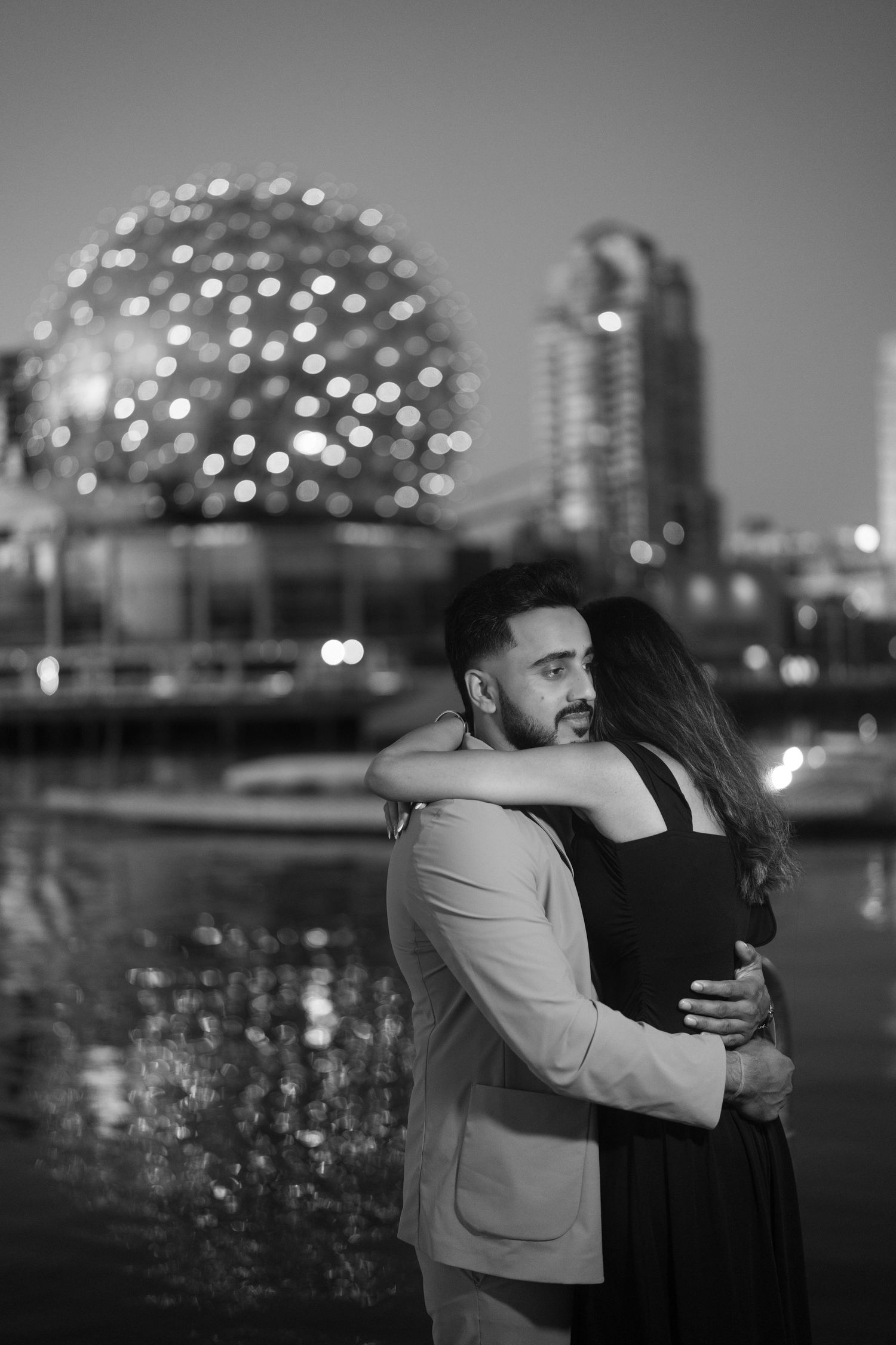 A romantic nighttime embrace against a cityscape backdrop with illuminated buildings and a glowing dome structure.