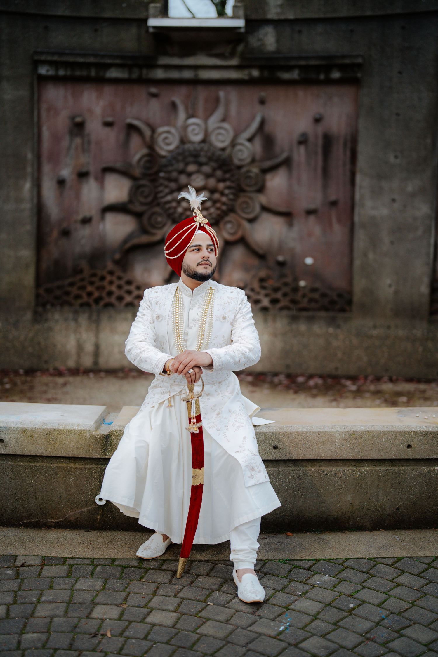 A person in white traditional attire and red turban poses on cobblestone ground against dark wall.