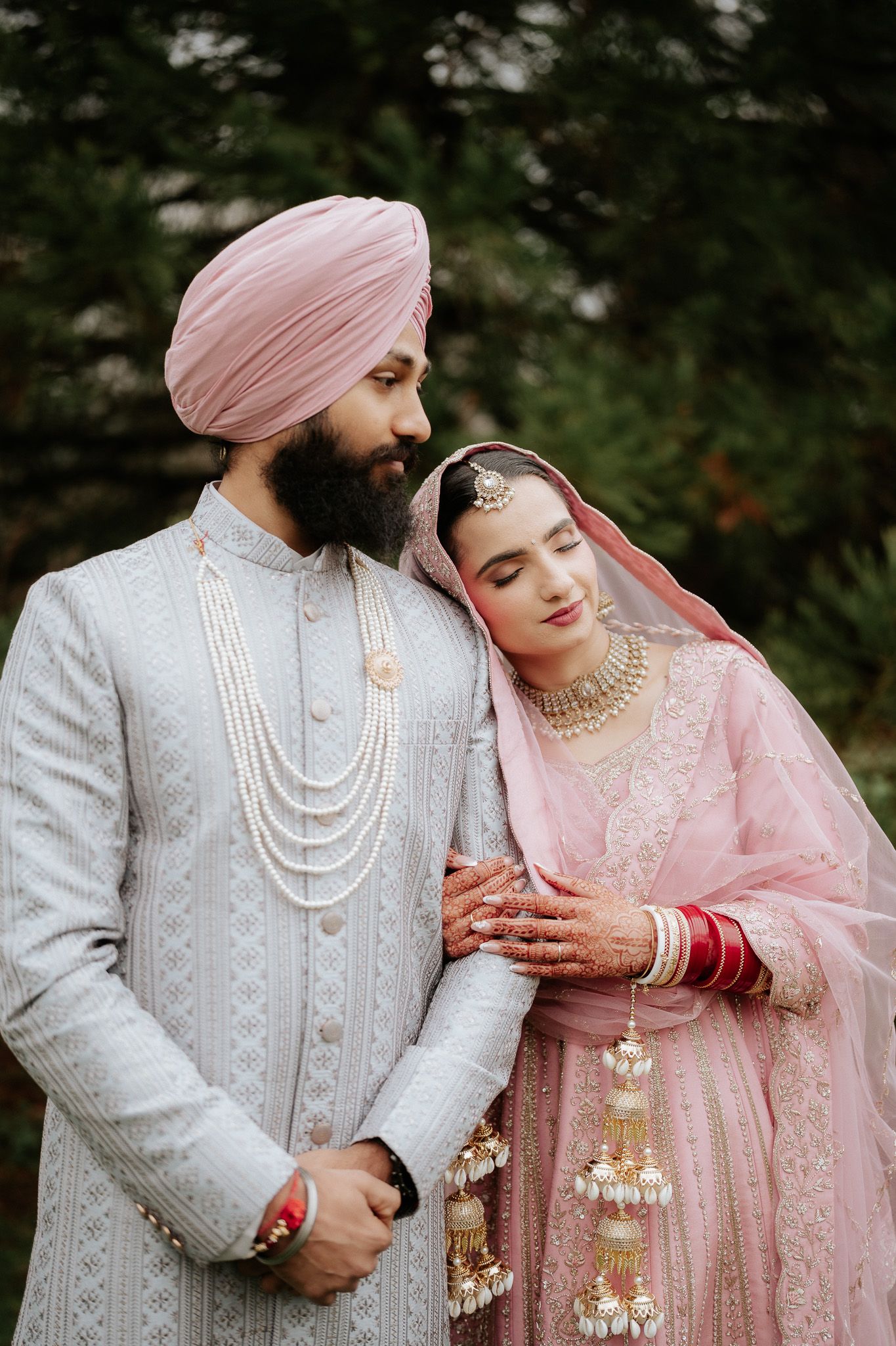 A romantic wedding couple in traditional Sikh attire with pink turban and pearl necklaces pose together in an outdoor setting.