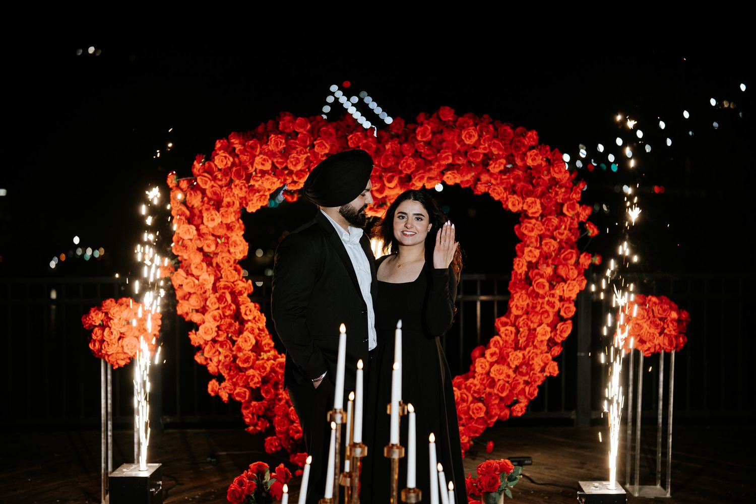 A romantic outdoor wedding ceremony setup with vibrant red floral arches and glowing candlelight at night.