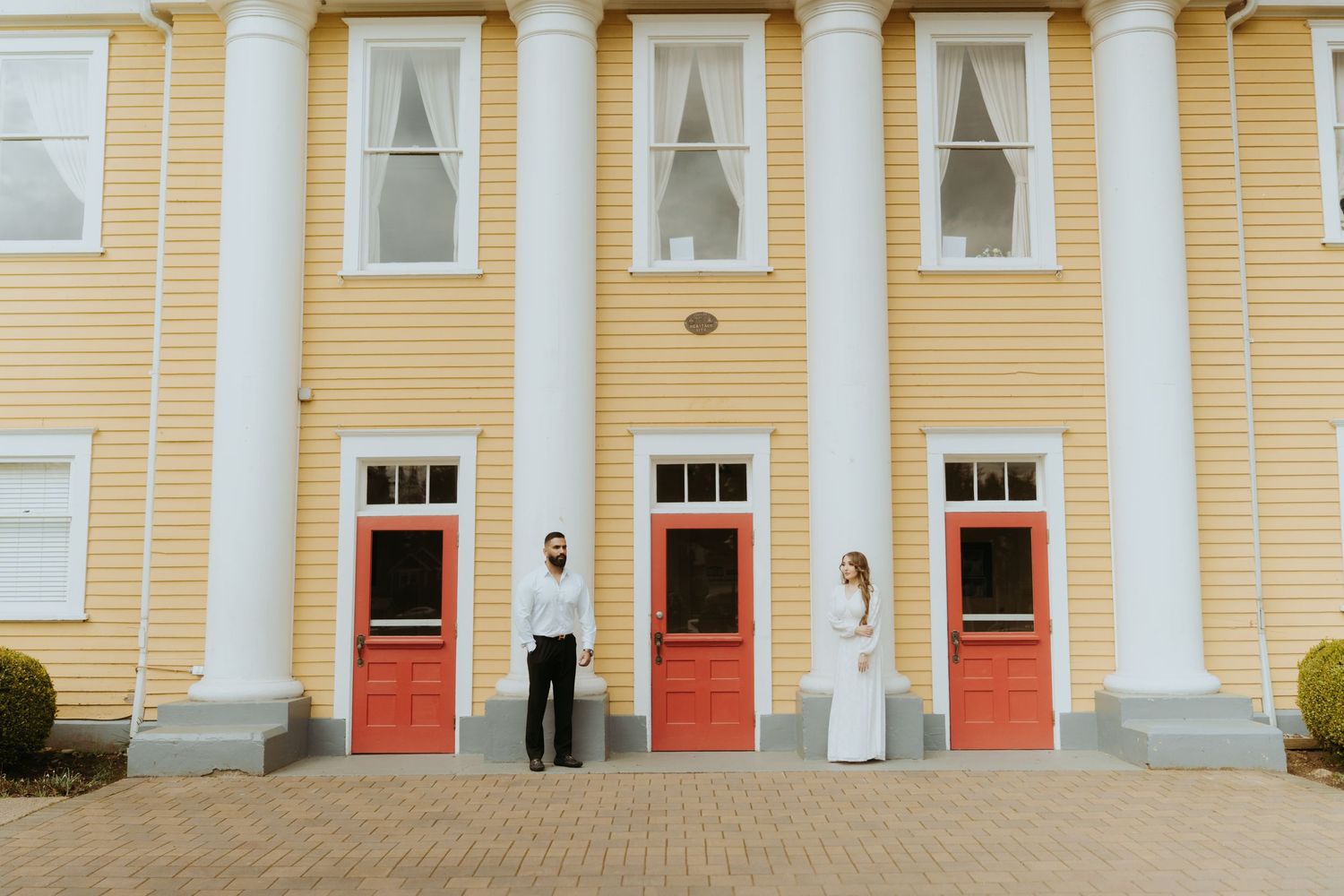 A couple stands in front of a yellow historic building with white columns and red doors.
