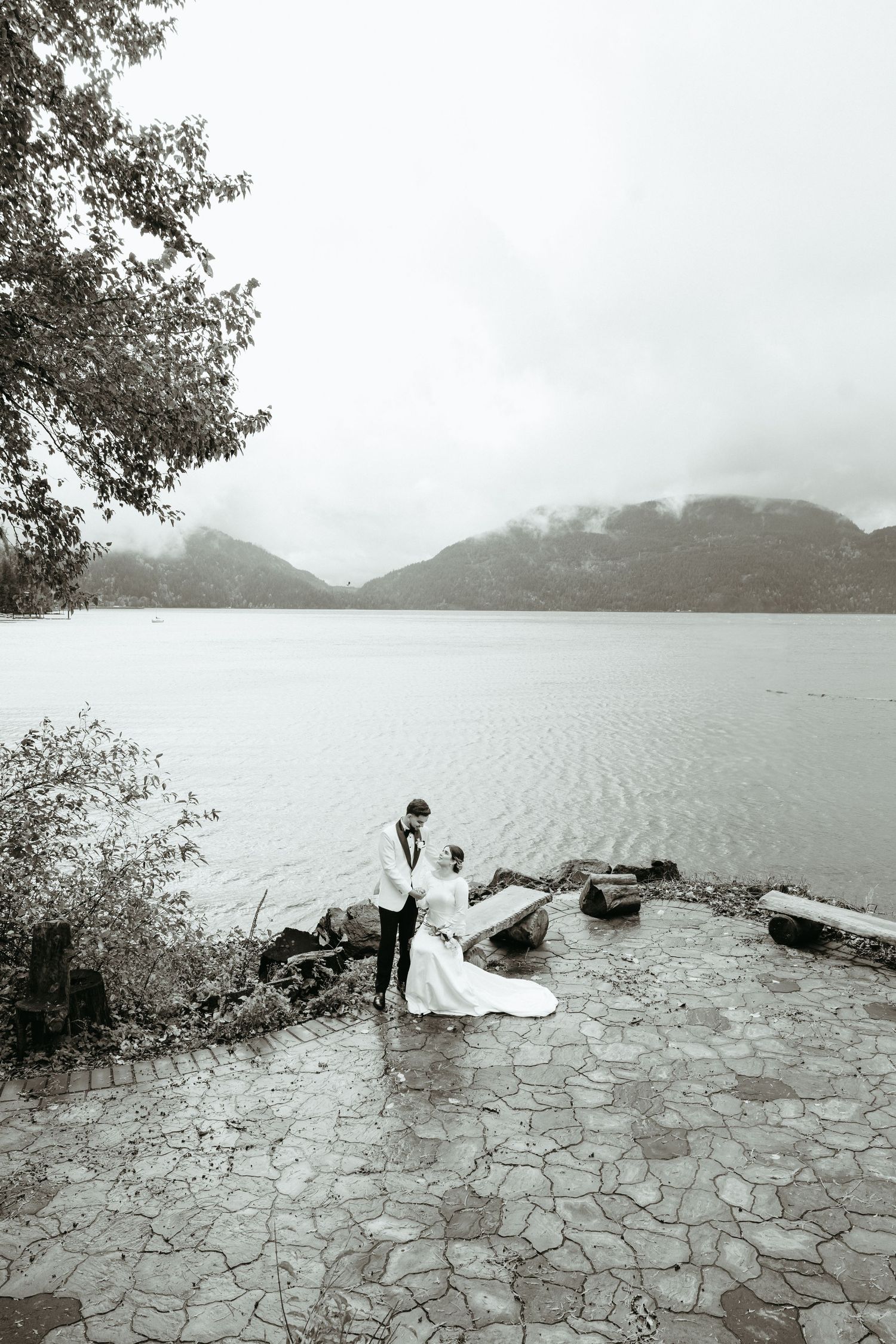 A serene black and white panoramic view of a lakeside stone path with mountains shrouded in mist in the background.