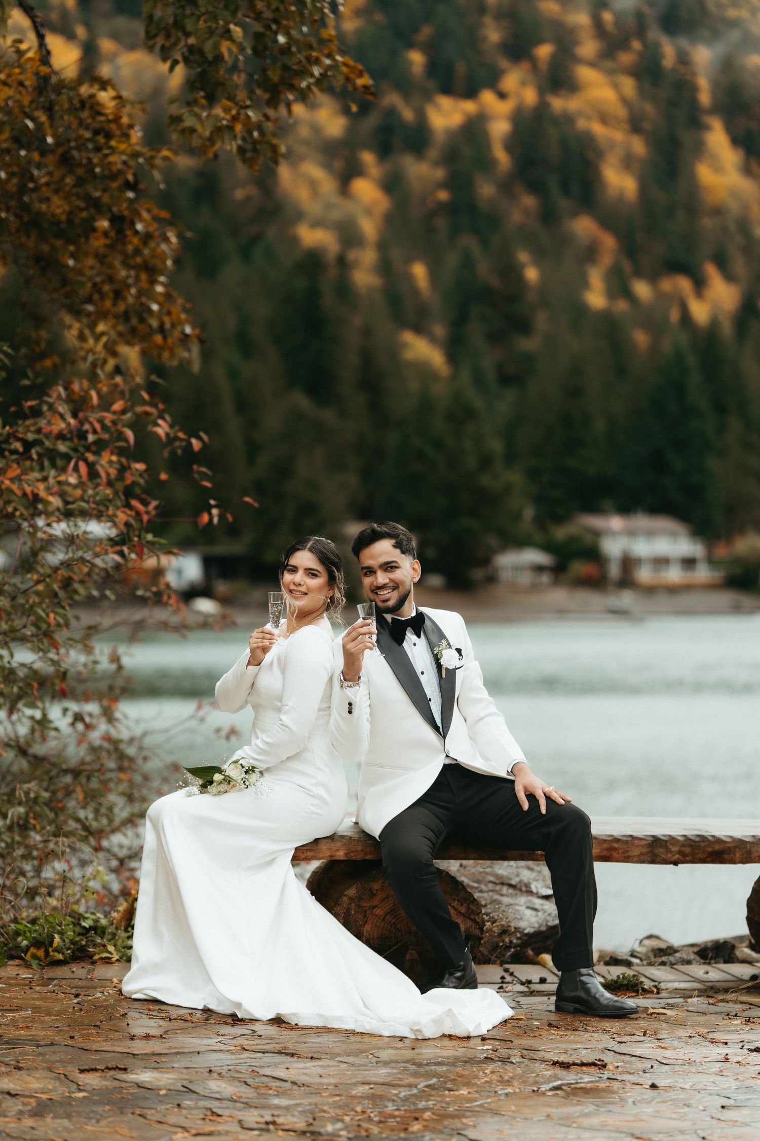 Couple in wedding attire sitting by a serene lake surrounded by autumn foliage during golden hour.