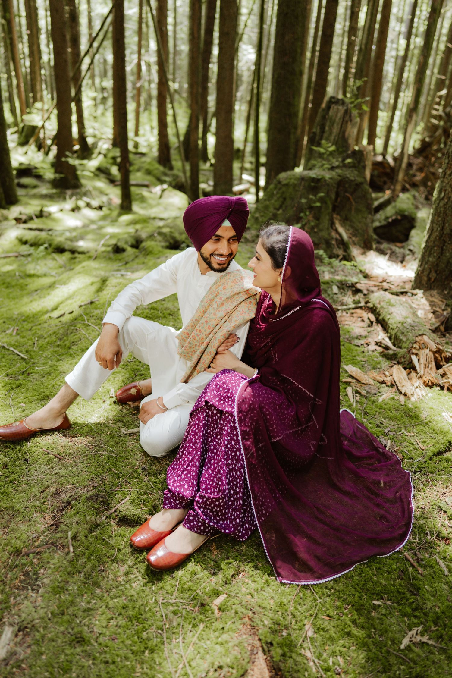 Sikh couple sitting on mossy forest ground, man in white kurta and purple turban smiling at woman.