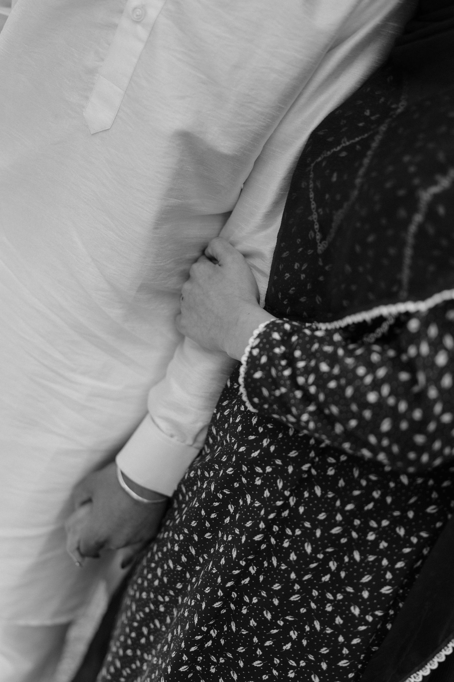 Black and white photo of a couple holding hands, bride in floral dress and groom in white shirt with bracelet.