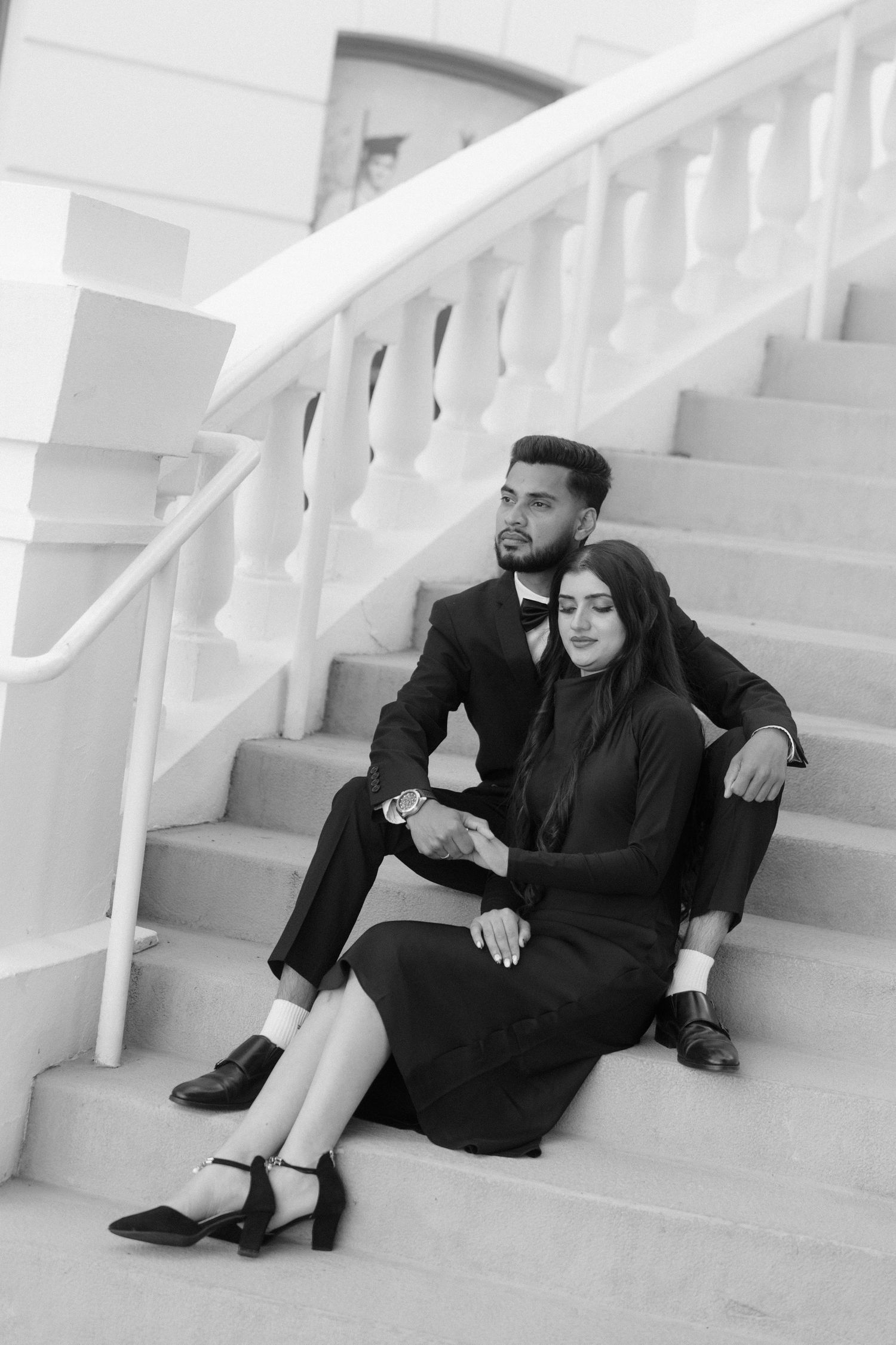 A couple dressed in matching black formal attire sits together on elegant white marble stairs.