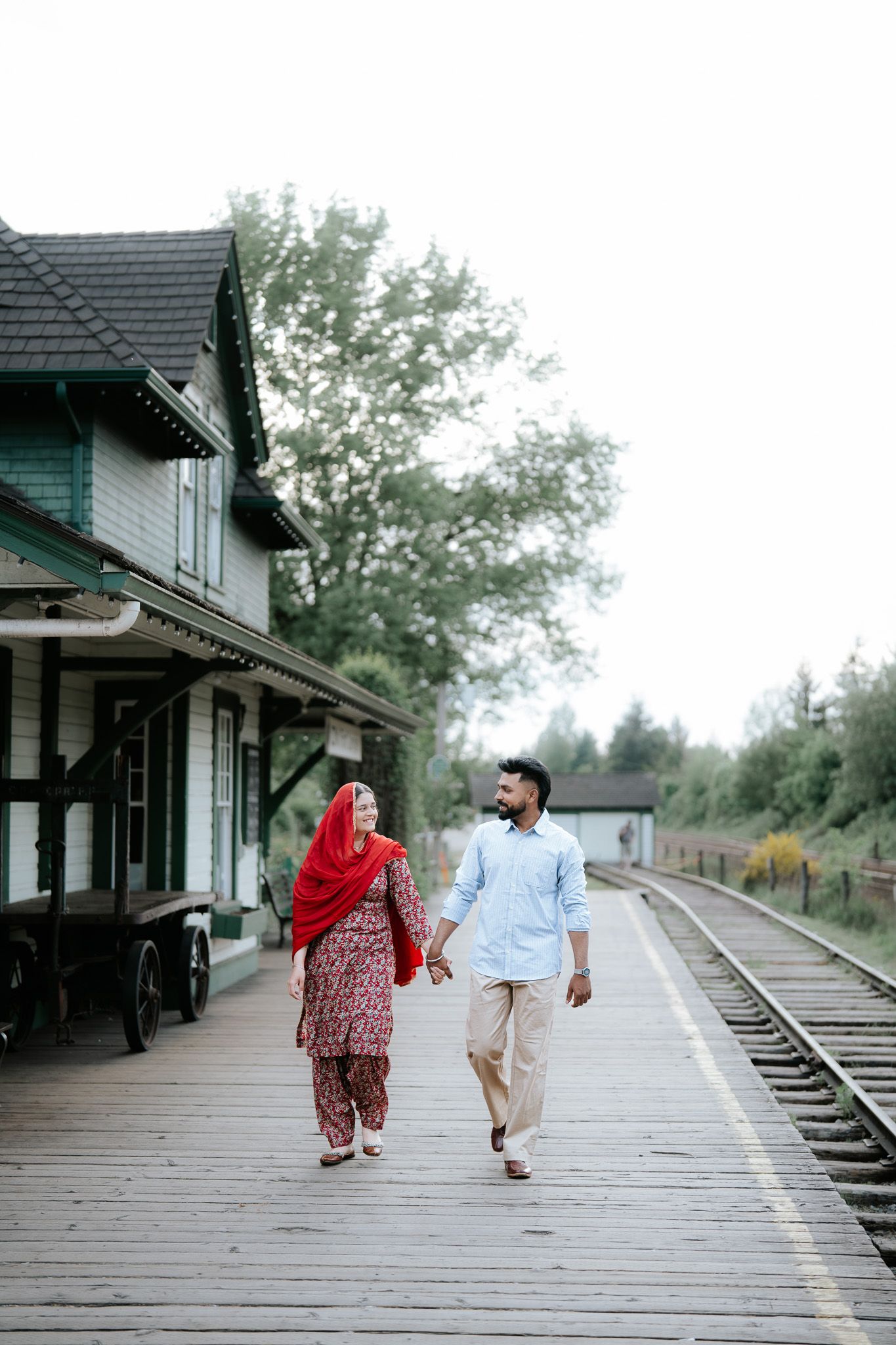 South Asian couple holding hands and walking along a wooden railway platform near a historic station.