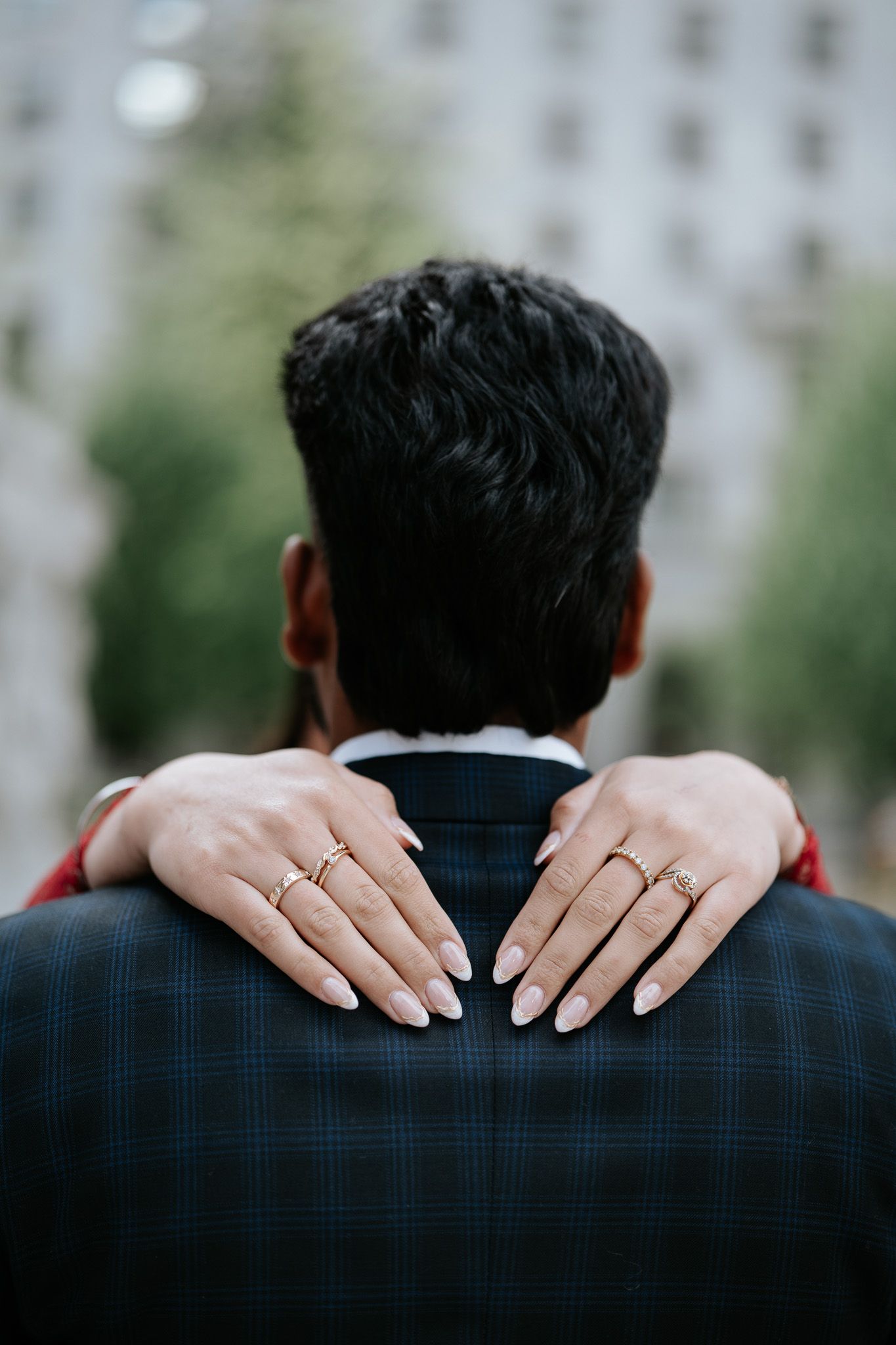 A woman's hands adorned with gold rings resting on a man's plaid suit jacket shoulders from behind.