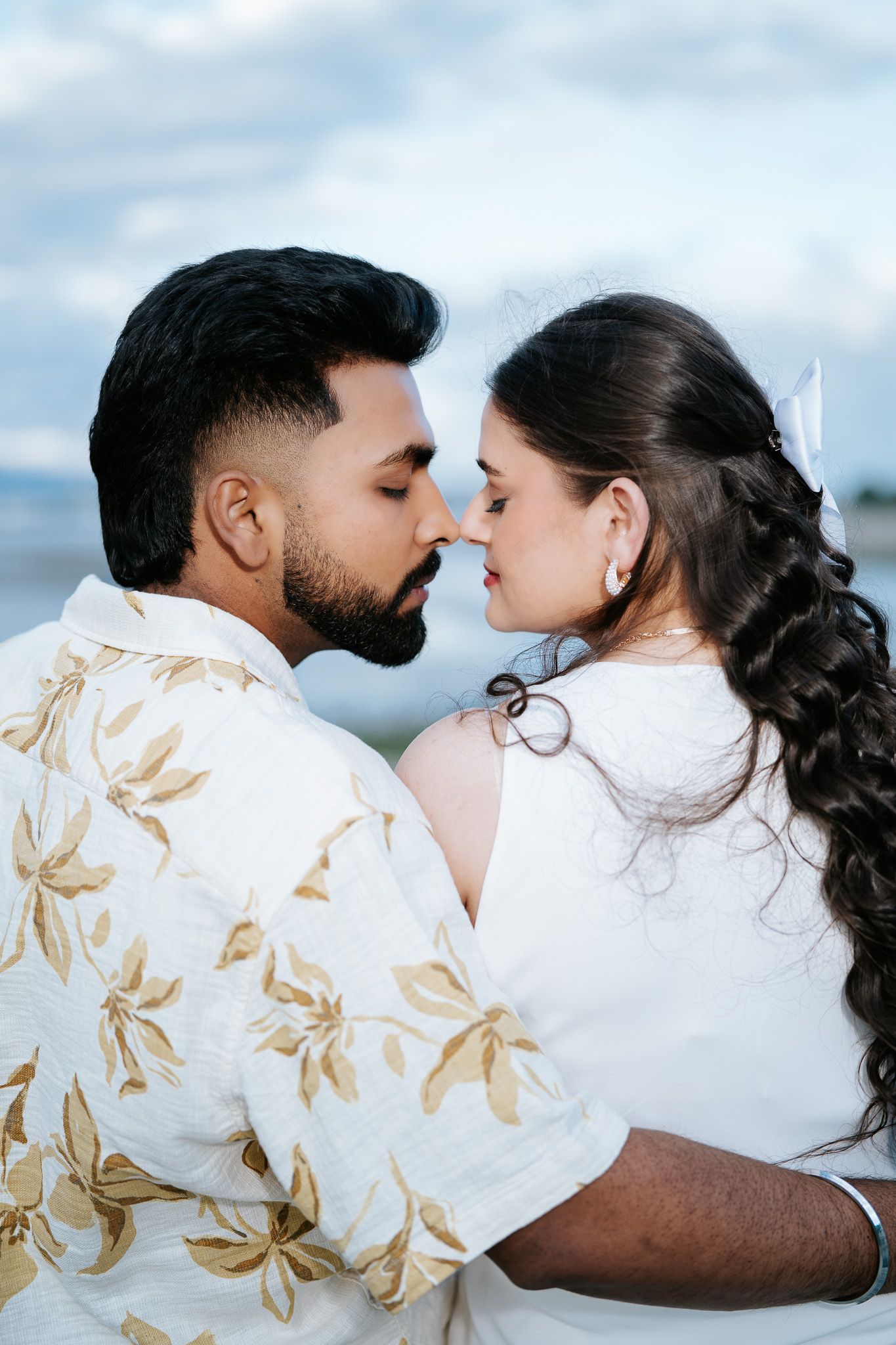 Romantic couple in white outfits sharing an intimate moment, nearly kissing against a cloudy sky backdrop.