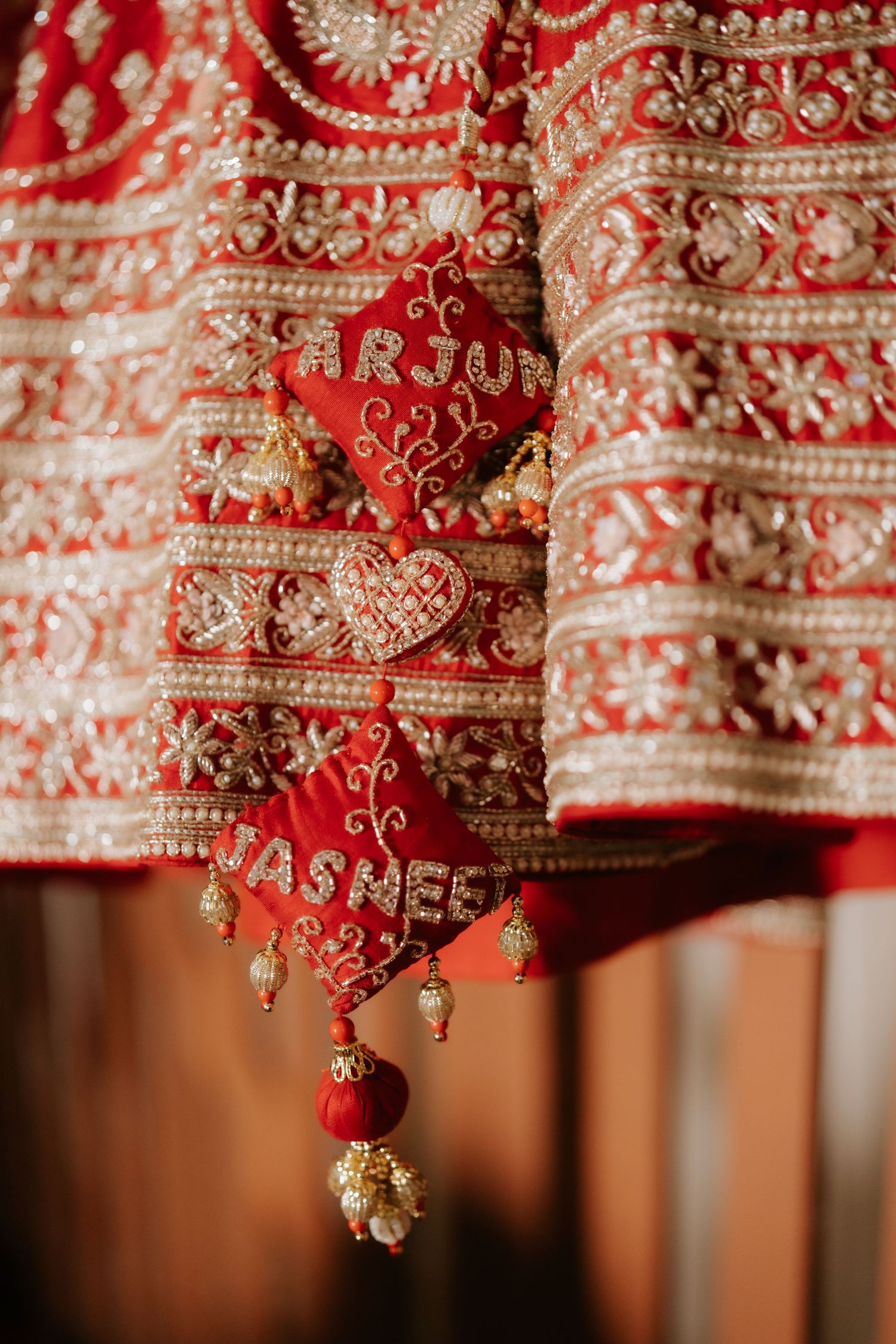 Red Indian bridal lehenga with gold embroidery and personalized name tags reading Arjun and Jashe with pearl tassels.