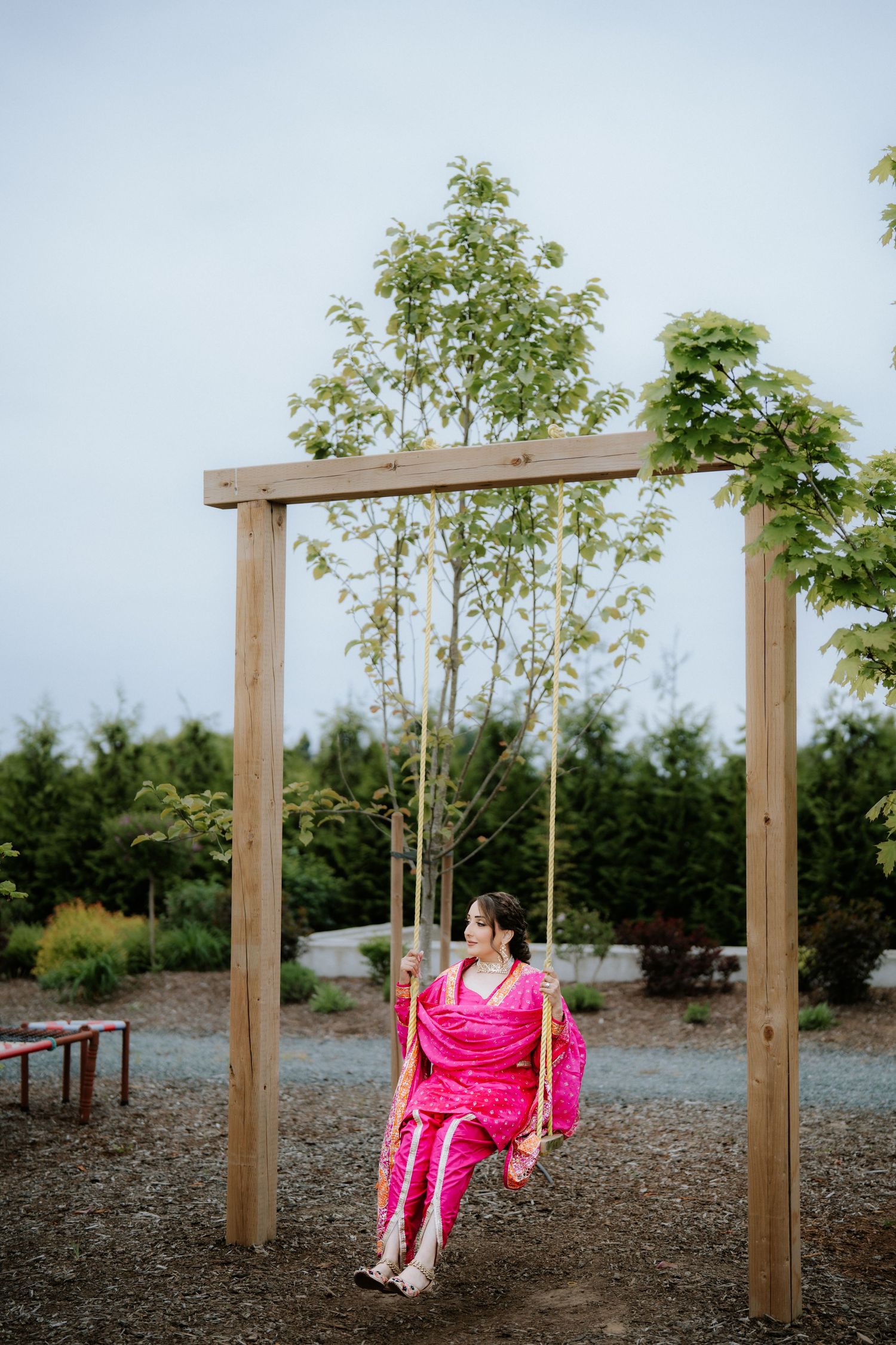 Woman in pink outfit sitting on a wooden swing surrounded by lush green trees in an outdoor garden setting.
