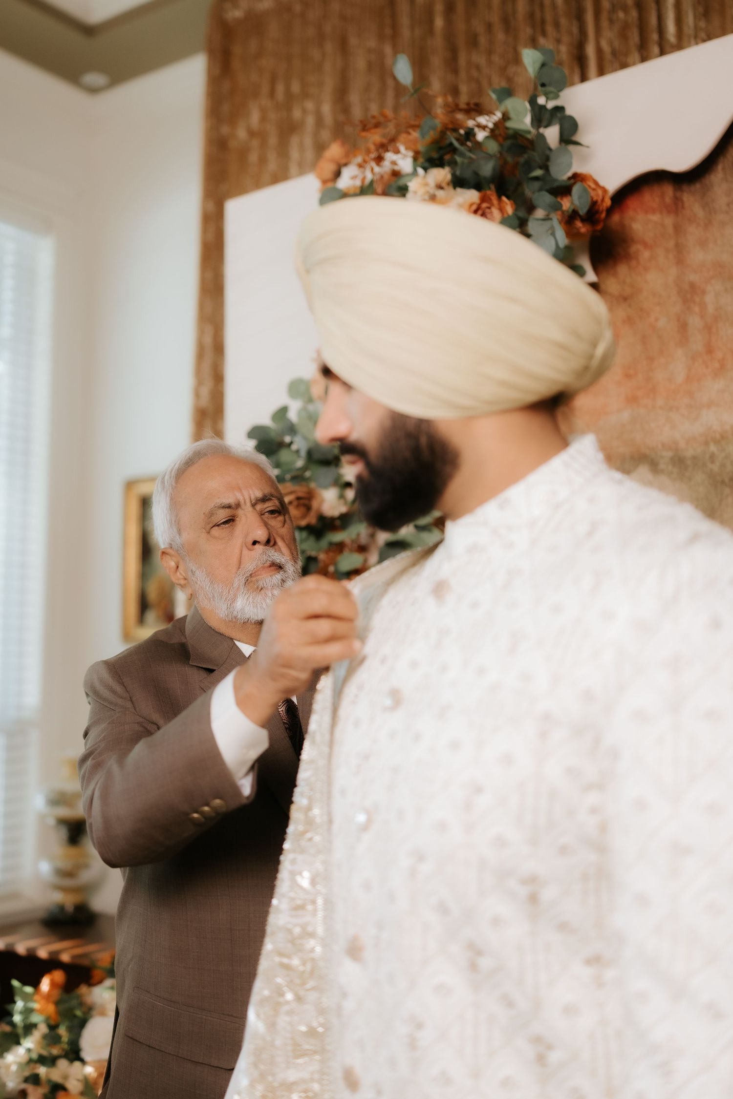 An elderly man in a brown suit adjusts the sherwani of a Sikh groom wearing a white turban at a wedding.