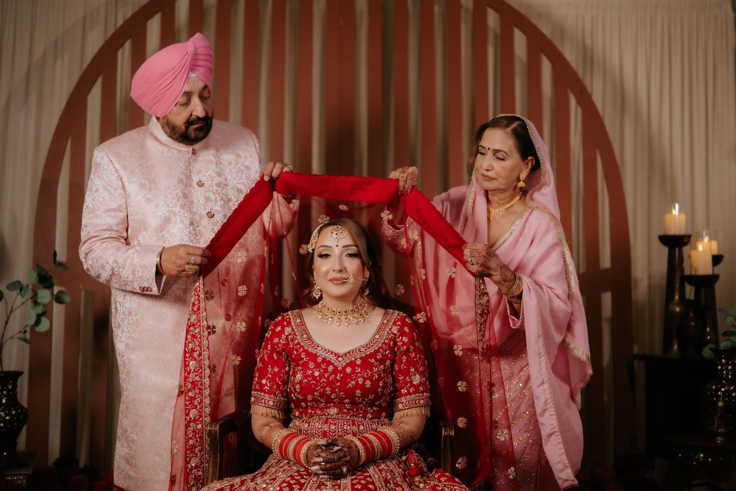 Sikh bride in red lehenga seated as family members hold red chunni during traditional wedding ceremony.