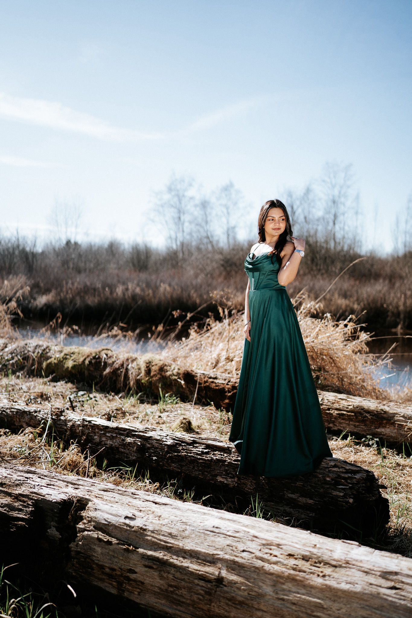 A woman in an emerald green dress poses gracefully on a fallen log in a winter landscape.