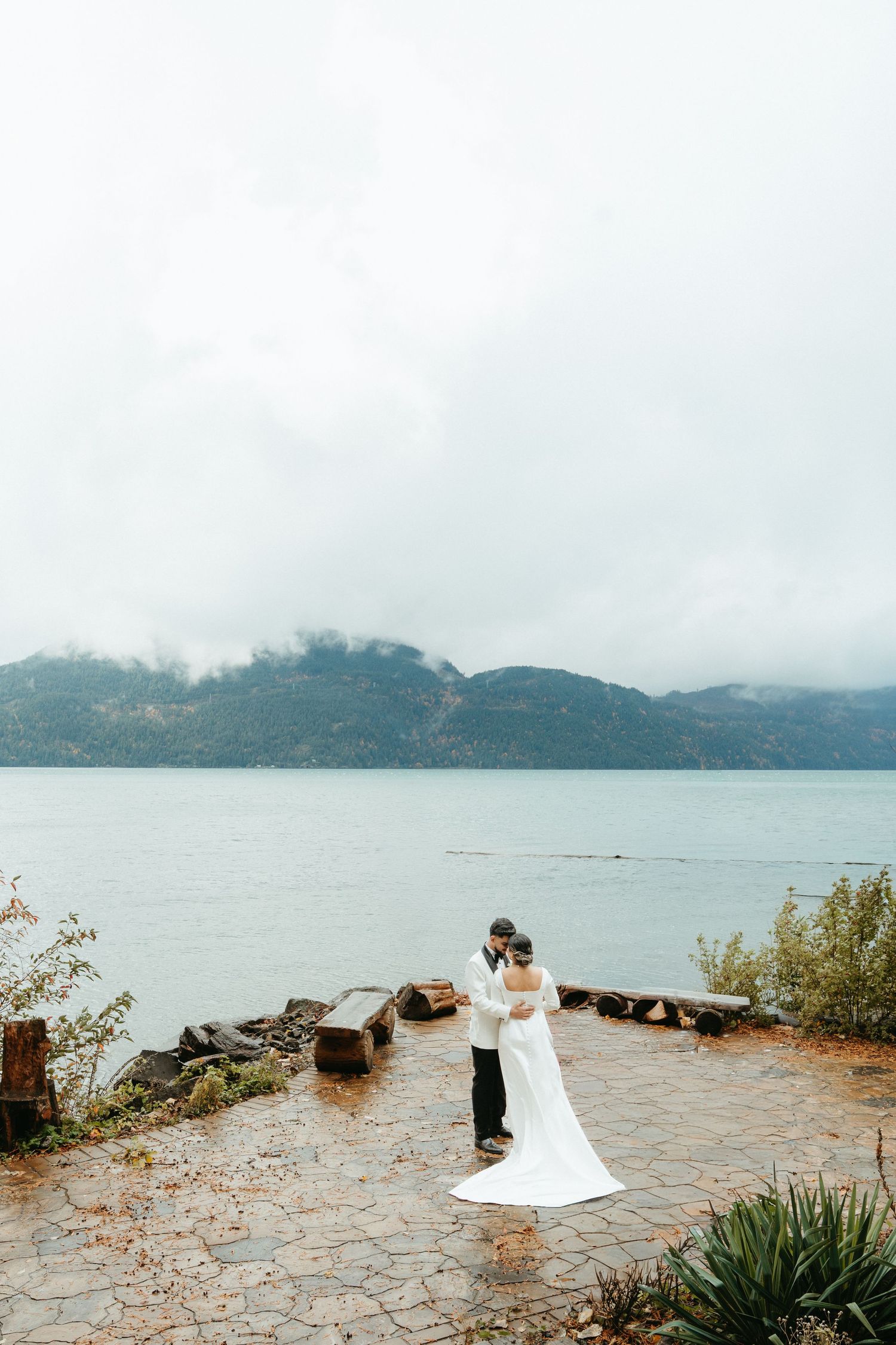 Couple embraces on stone pathway overlooking misty mountain lake on overcast day.
