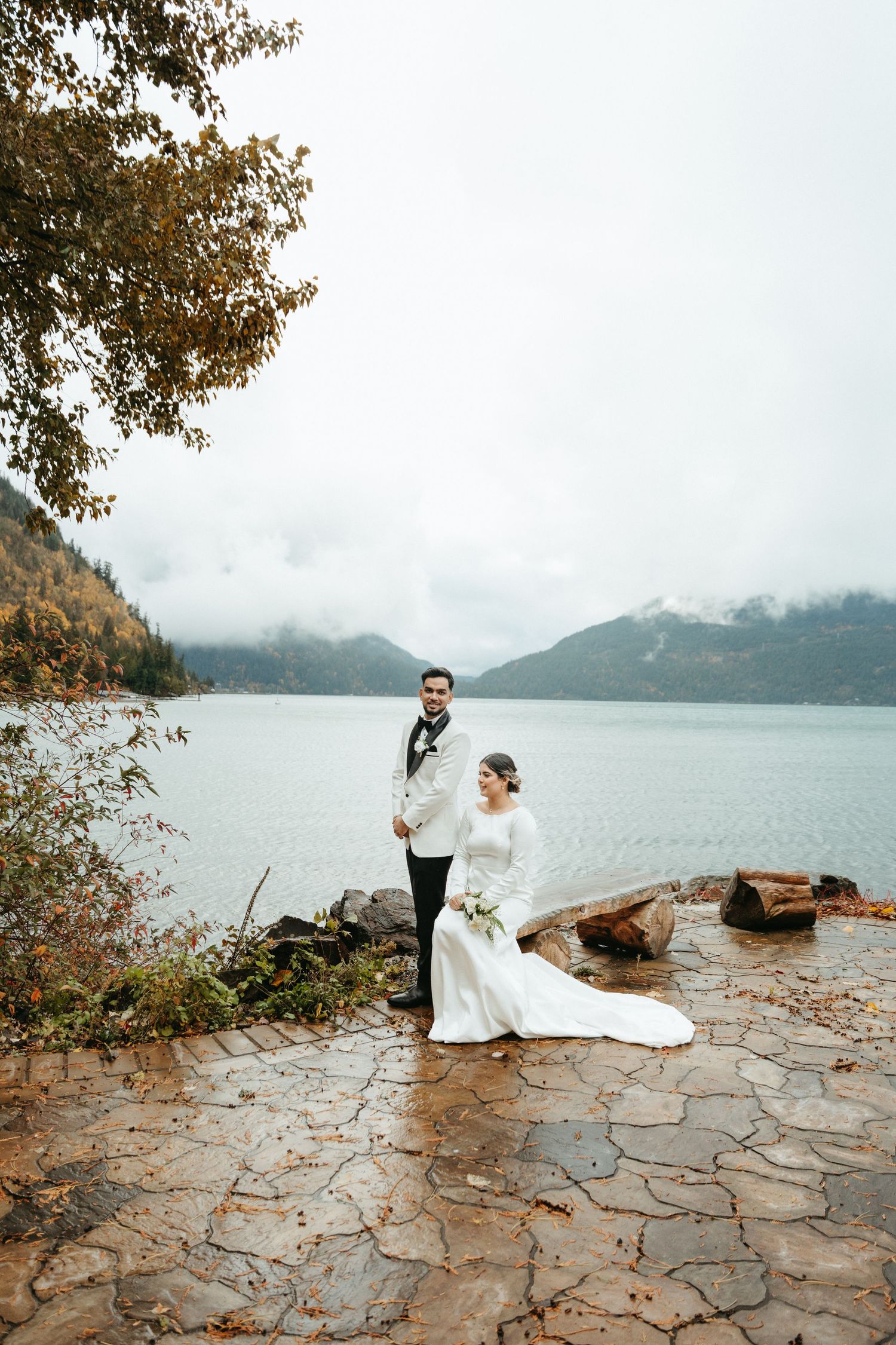 A couple in wedding attire poses on a stone path overlooking a misty mountain lake on an autumn day.