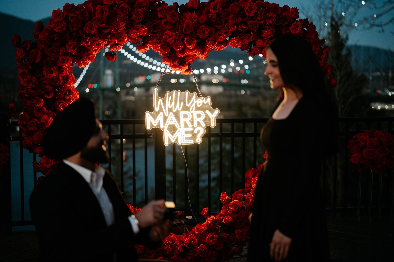 A romantic marriage proposal scene with glowing 'Marry' sign and red floral arch against an evening sky.