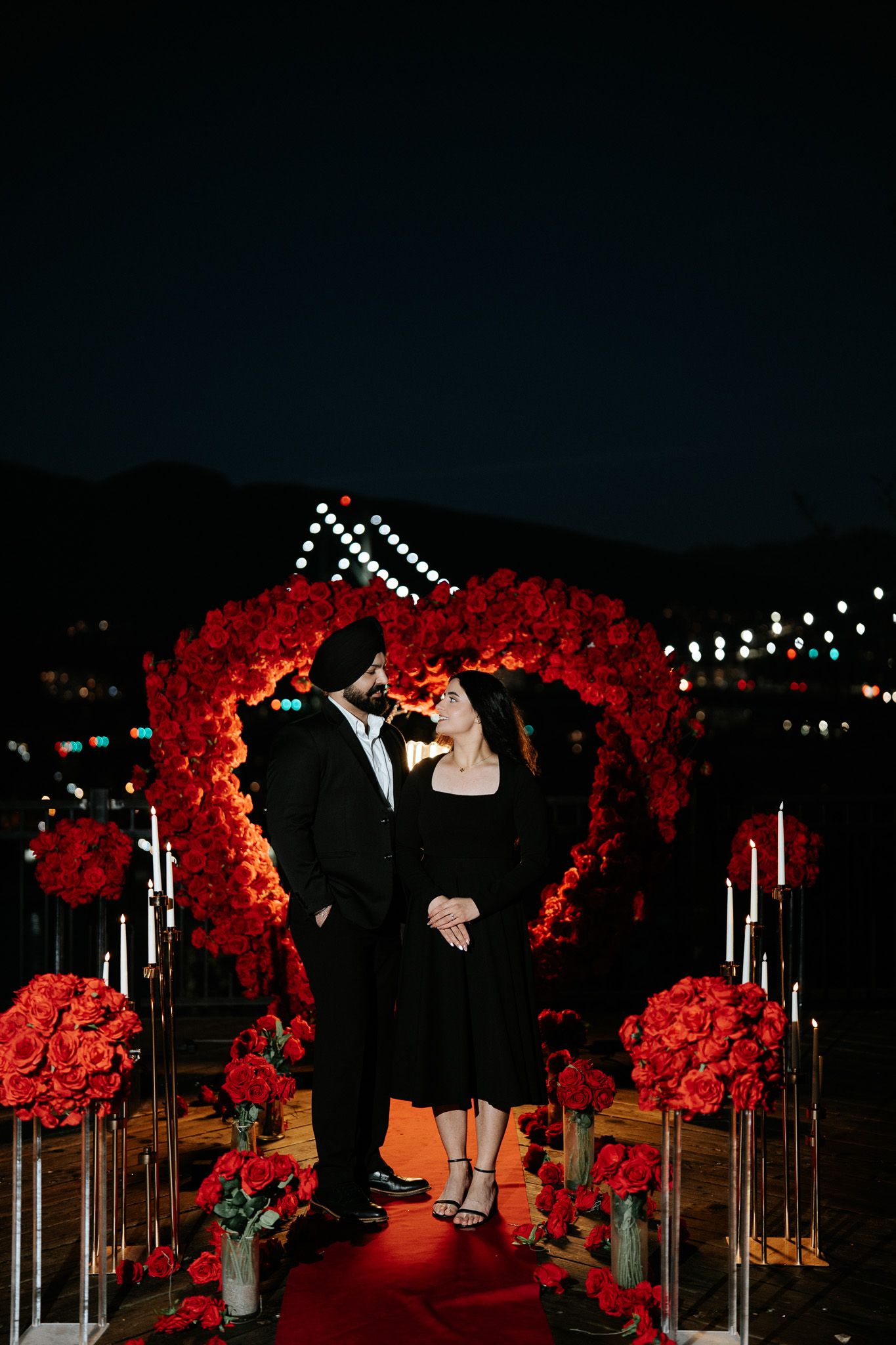 A romantic proposal setup at night with a heart-shaped red floral arch, candles, and string lights against a dark sky.