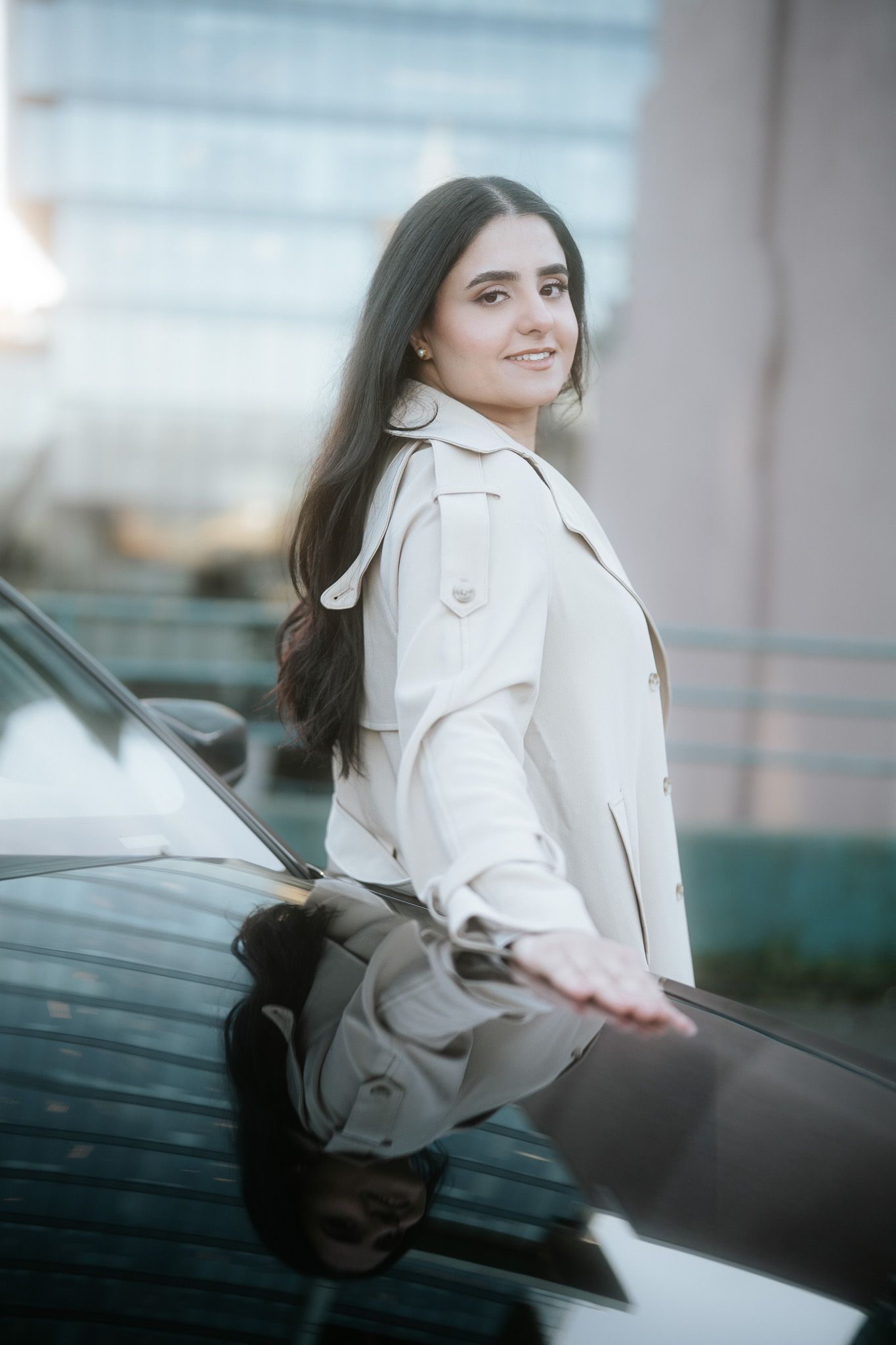 Someone in a beige trench coat poses with a black car in an urban setting with soft lighting.