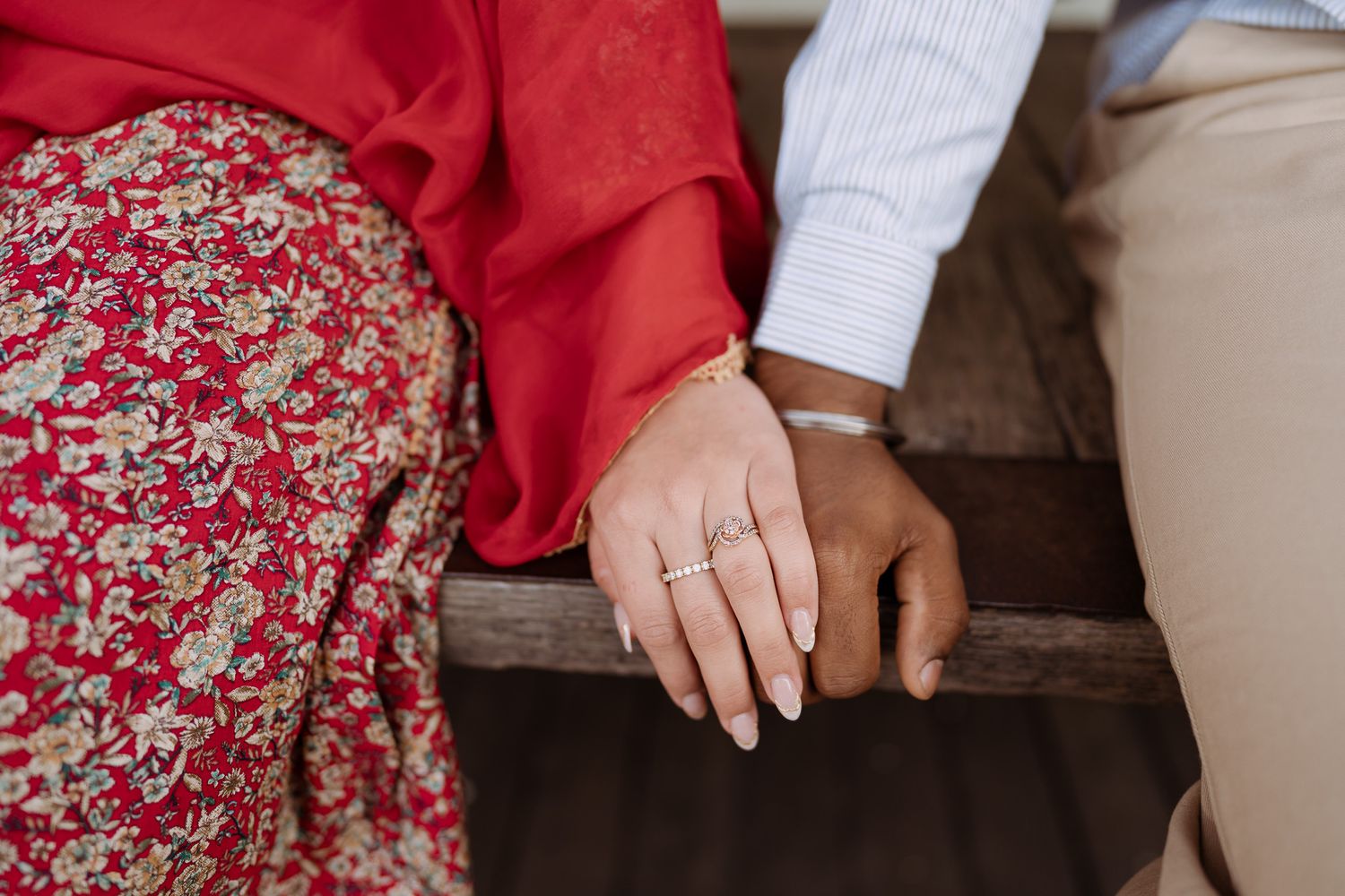 Close-up of a couple holding hands on a wooden bench, woman wearing gold bangles and rings in red floral attire.