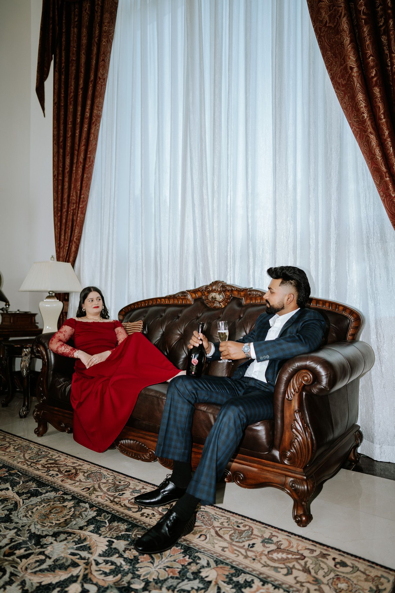 Elegant couple in formal attire relaxing on a leather sofa with champagne in a luxurious room.