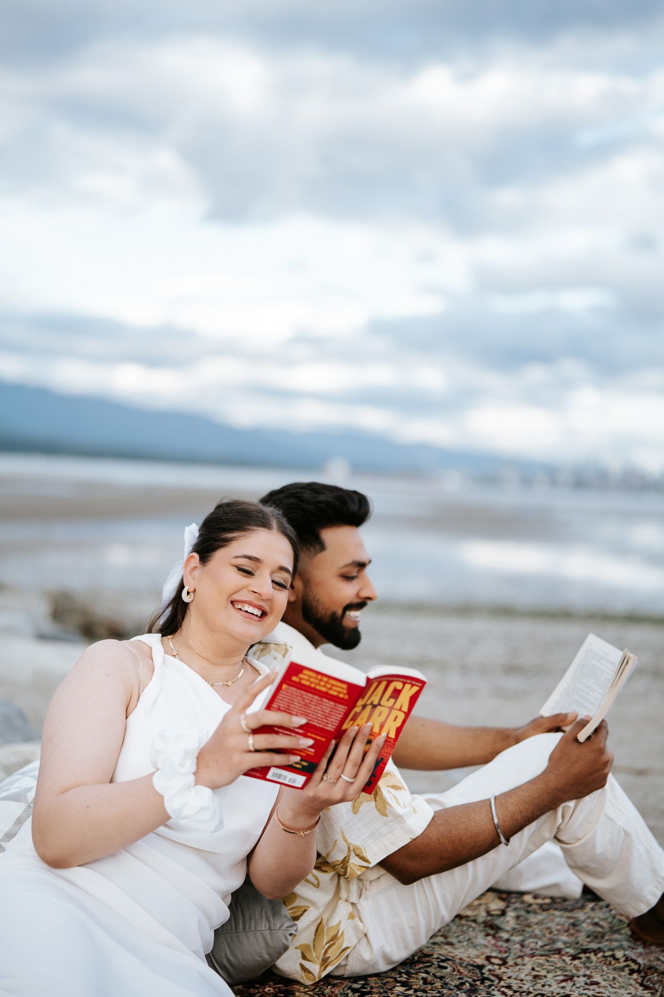 Happy couple reading Jack Carr book together on rocky beach, both dressed in white, overcast sky behind.