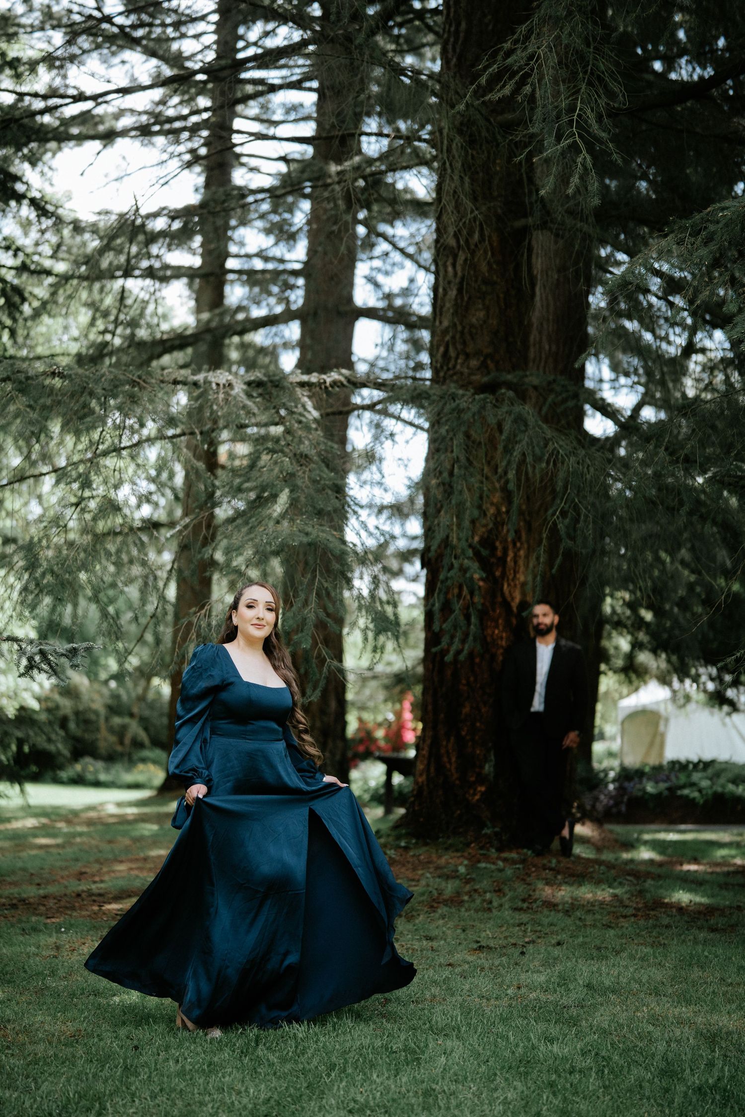 Woman in teal gown twirling in forest with man standing by trees in background.