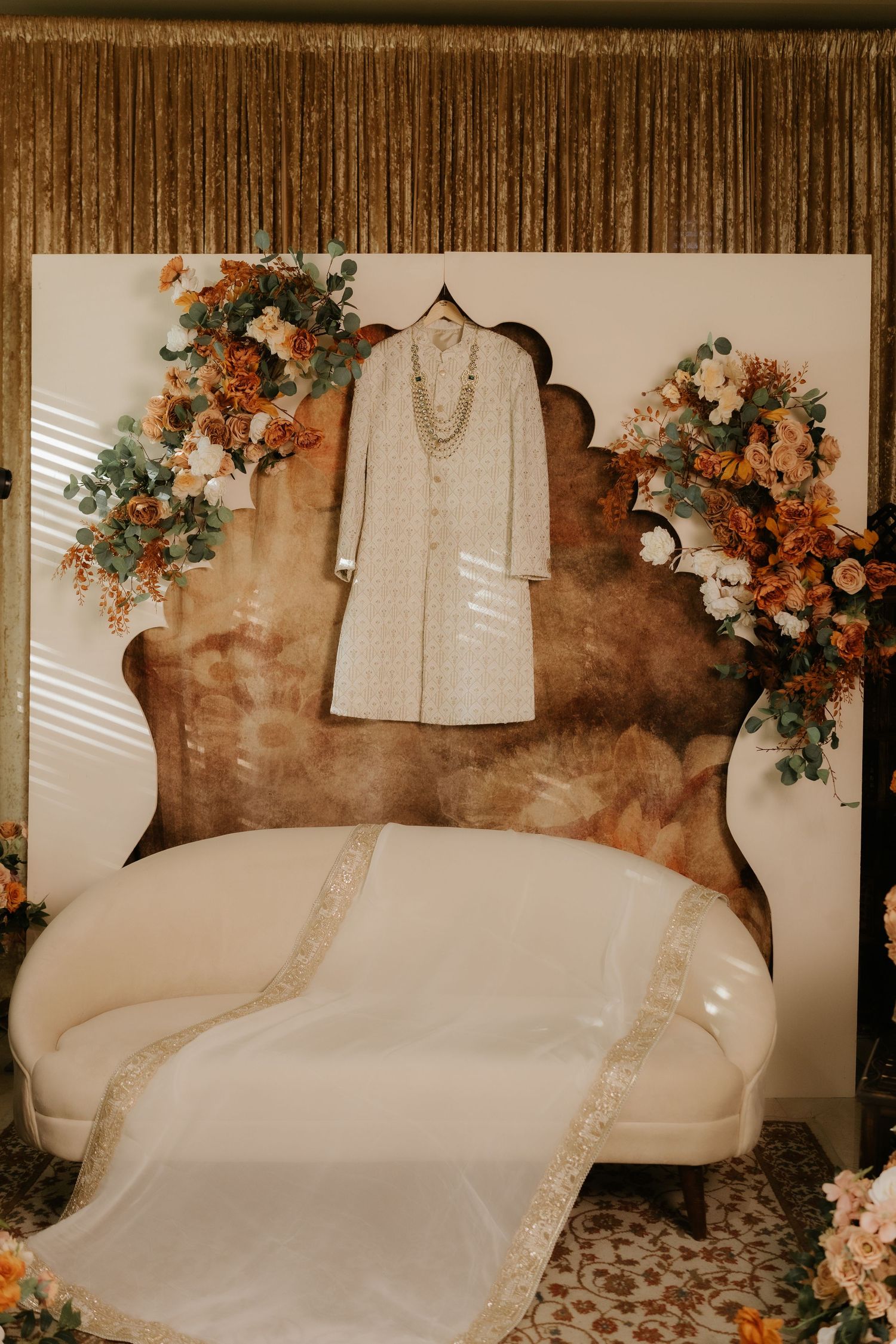 Elegant cream sherwani displayed on a hanger above a white sofa, surrounded by autumn floral arrangements in warm tones.