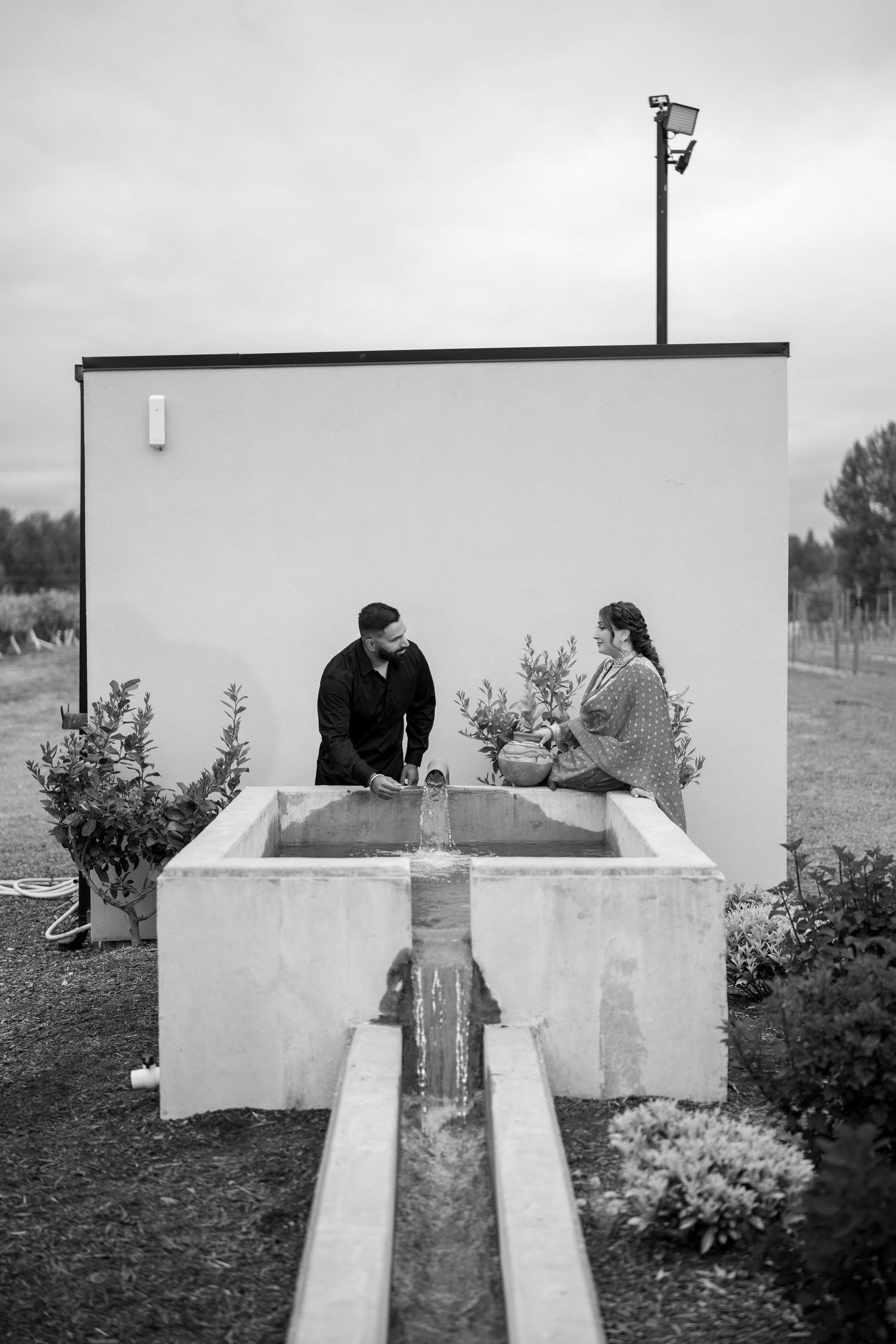 Black and white photo of a couple standing at an outdoor concrete water fountain feature near a white building.