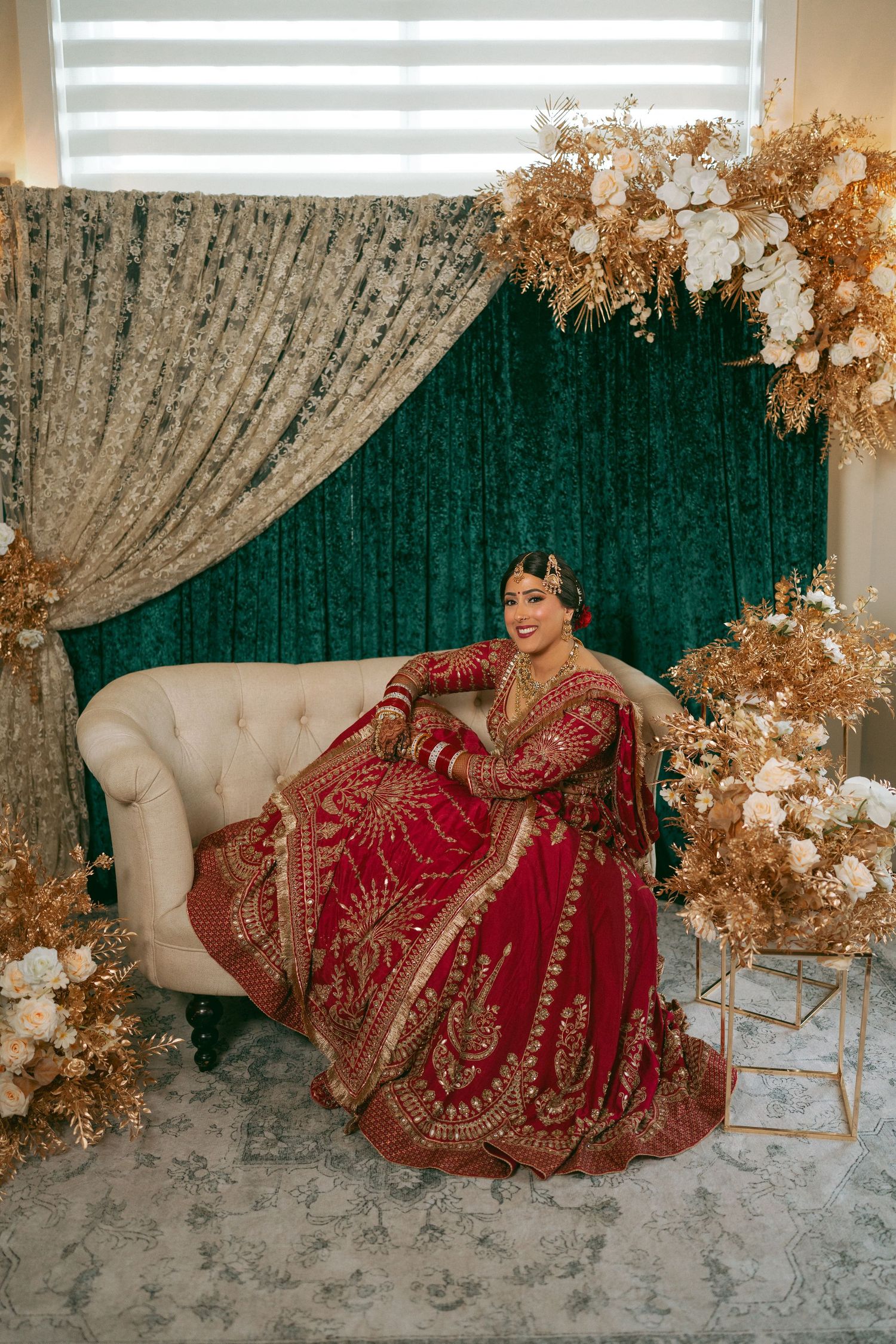 Bride in red and gold embroidered lehenga sits on cream sofa with green velvet backdrop and golden floral decor.