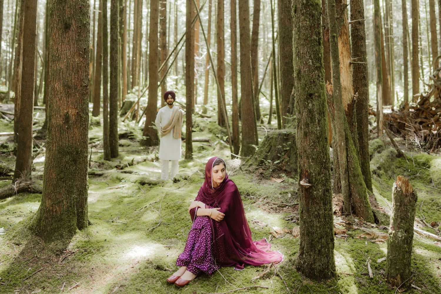Woman in purple dress sits by forest tree while man stands among tall woodland trees on mossy ground.