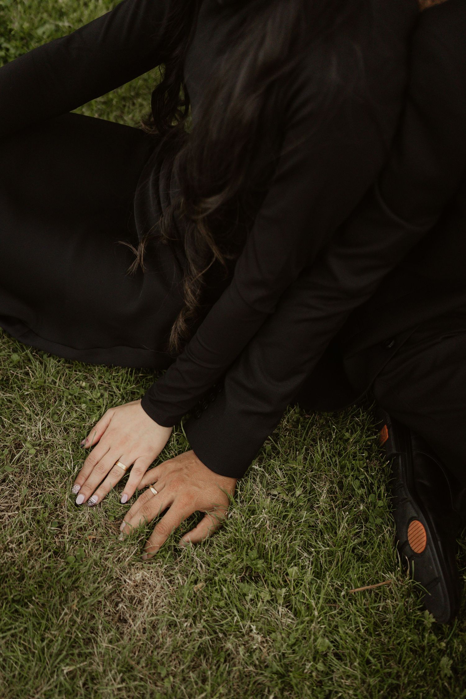 A woman in black sits on grass, displaying an engagement ring on her hand beside a man's shoe.