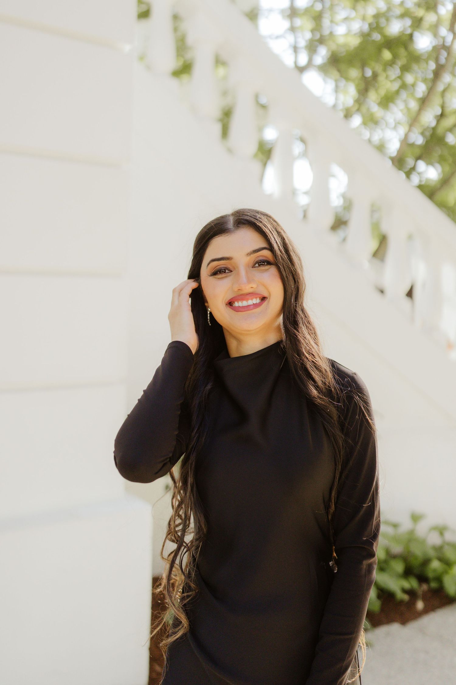 Smiling young woman with long dark hair in a black turtleneck dress posing near white balustrade outdoors.