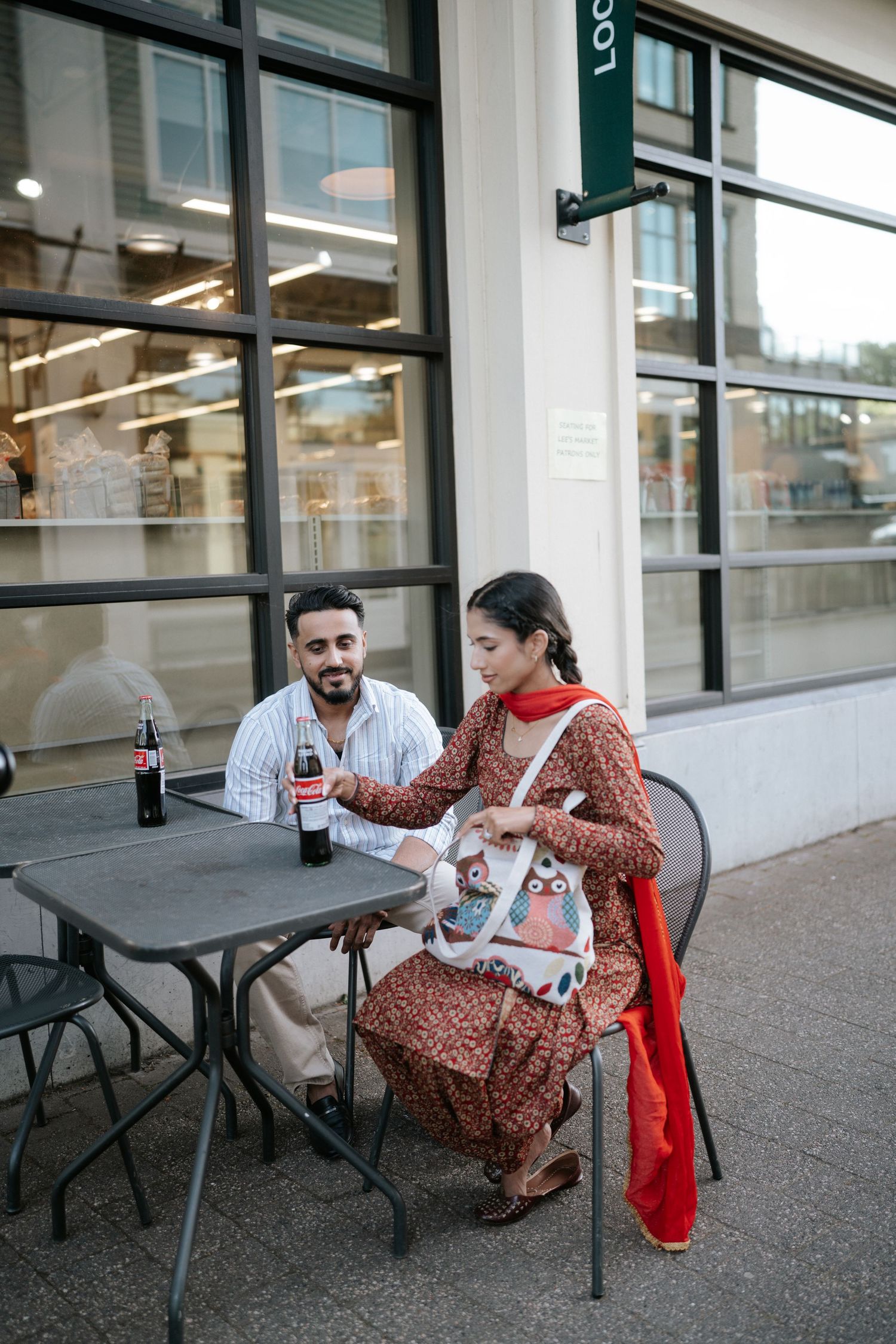 A couple shares a lighthearted moment while sitting at outdoor tables near a storefront window.
