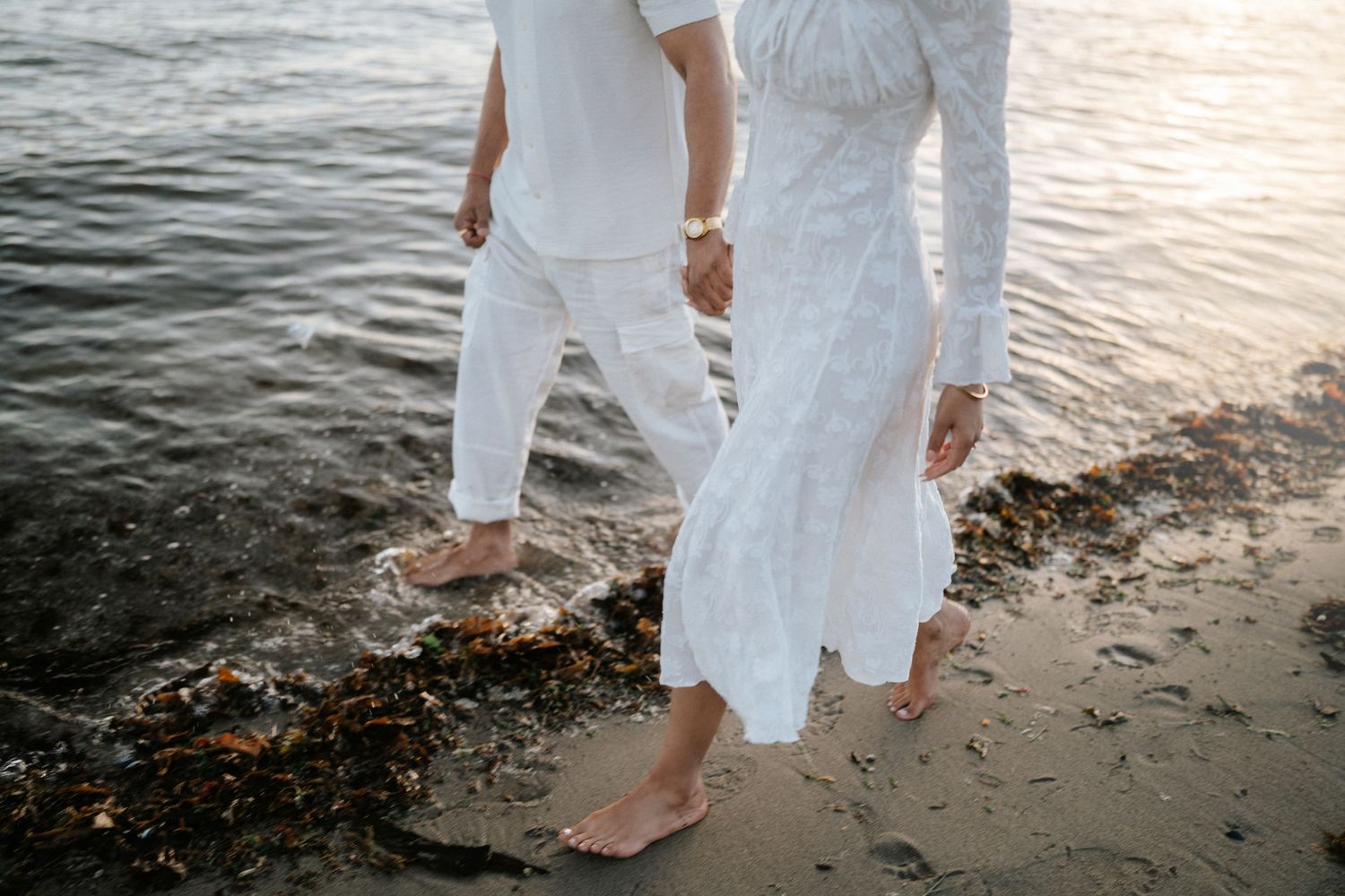 Person in flowing white dress walks barefoot along beach shoreline at sunset creating reflective silhouette on wet sand.