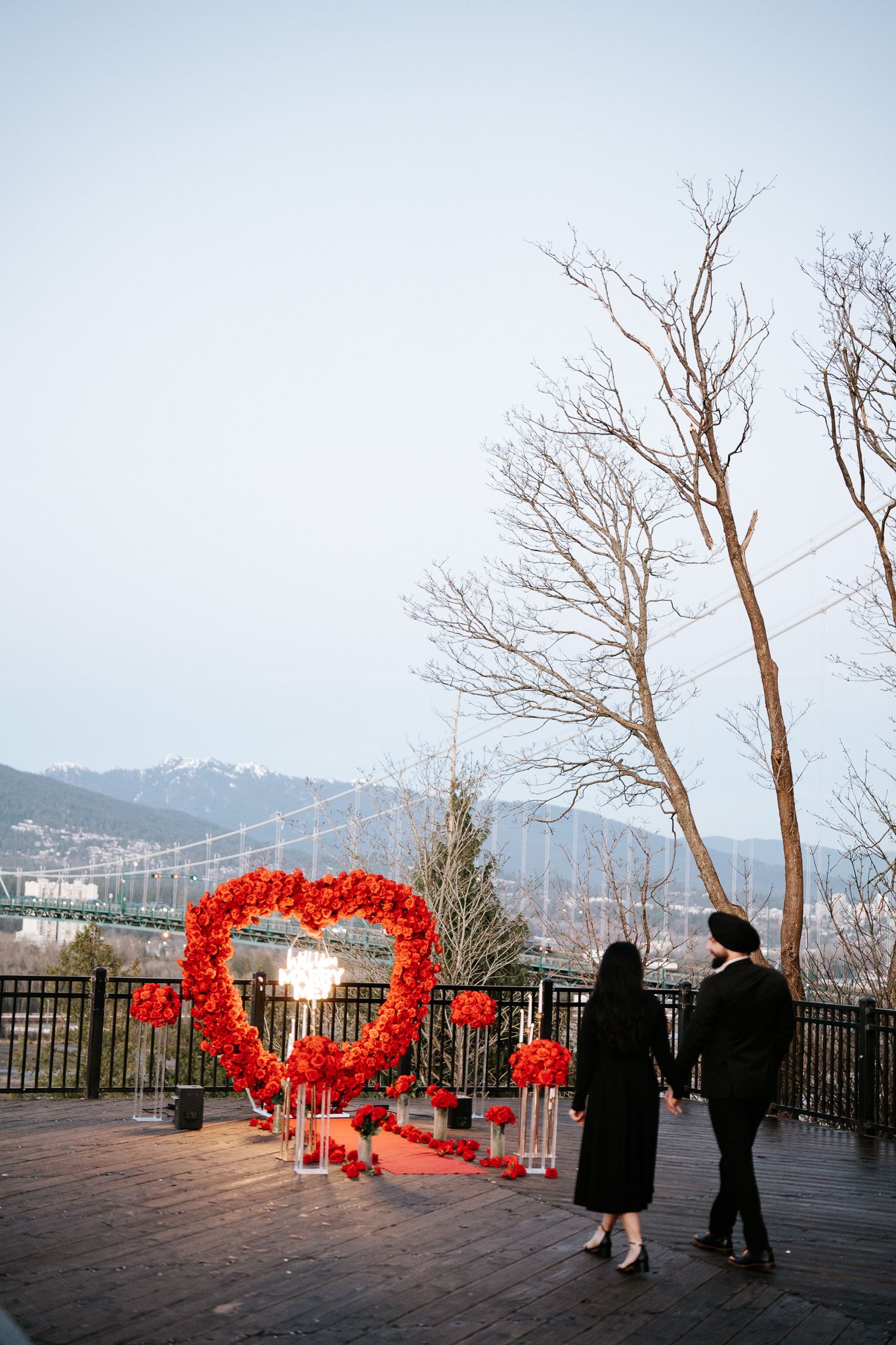 Couple stands near large red heart-shaped art installation on outdoor viewing deck with city view.